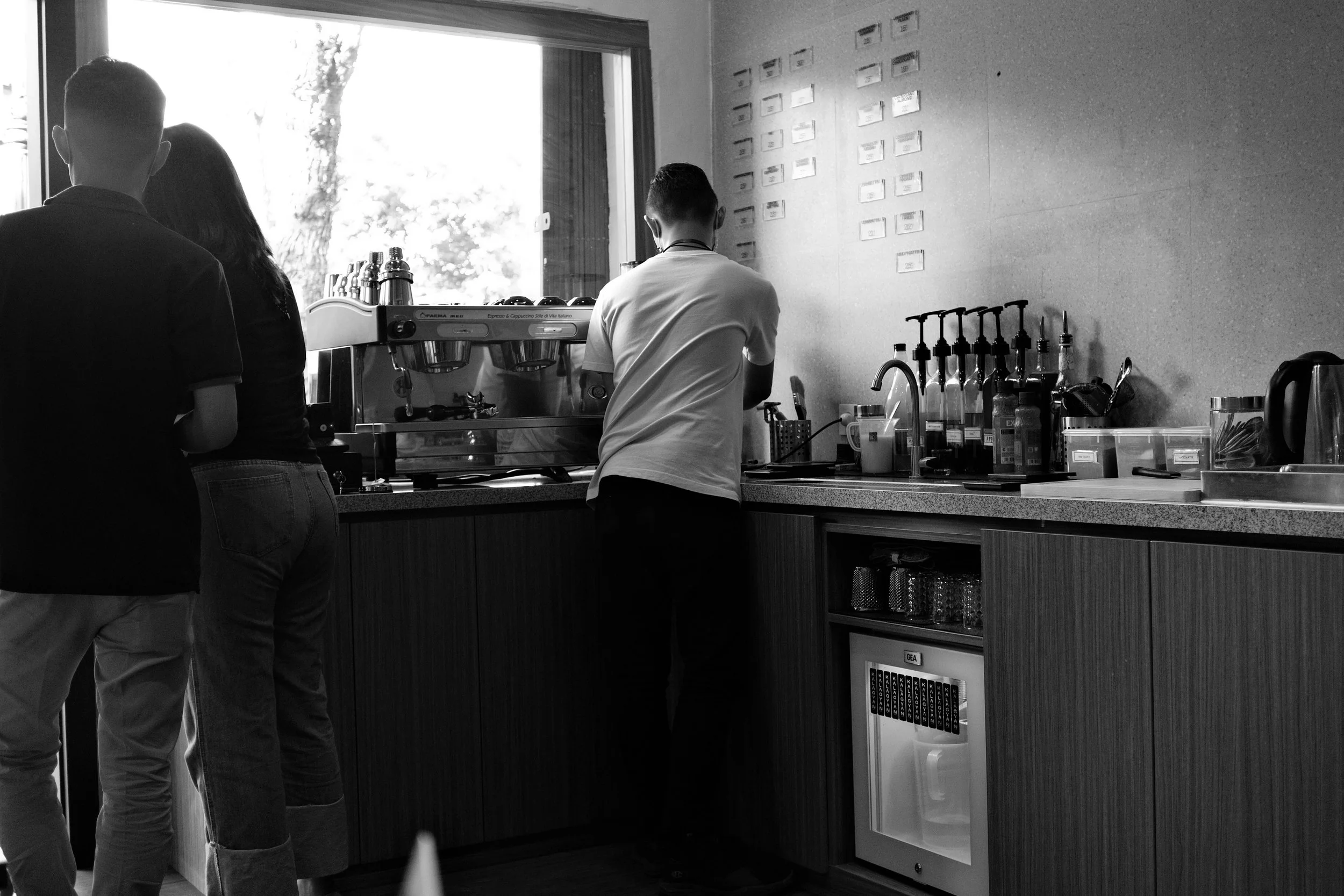 Three people inside a café or coffee shop, near a window. One person is behind the counter, operating an espresso machine. Two other people are standing in front of the counter, talking. The counter has various bottles, utensils, and a small refrigerator underneath.