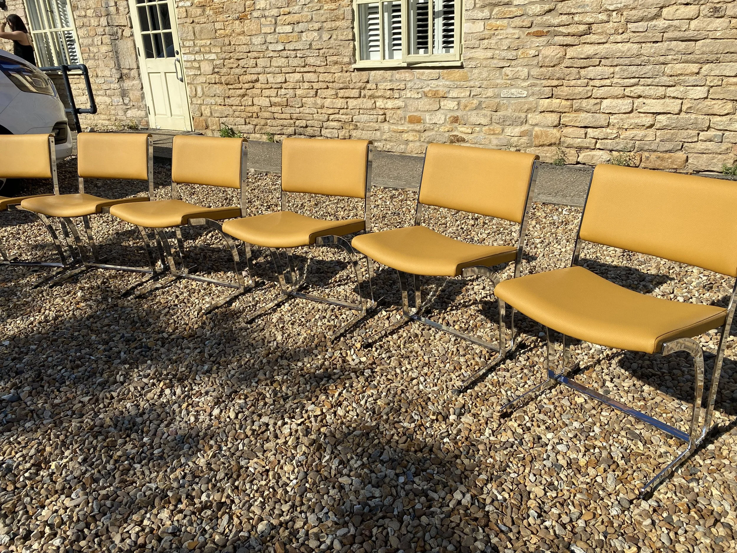 Six vintage yellow chairs with chrome frames lined up on a gravel surface outdoors, with a stone building and a white door in the background.