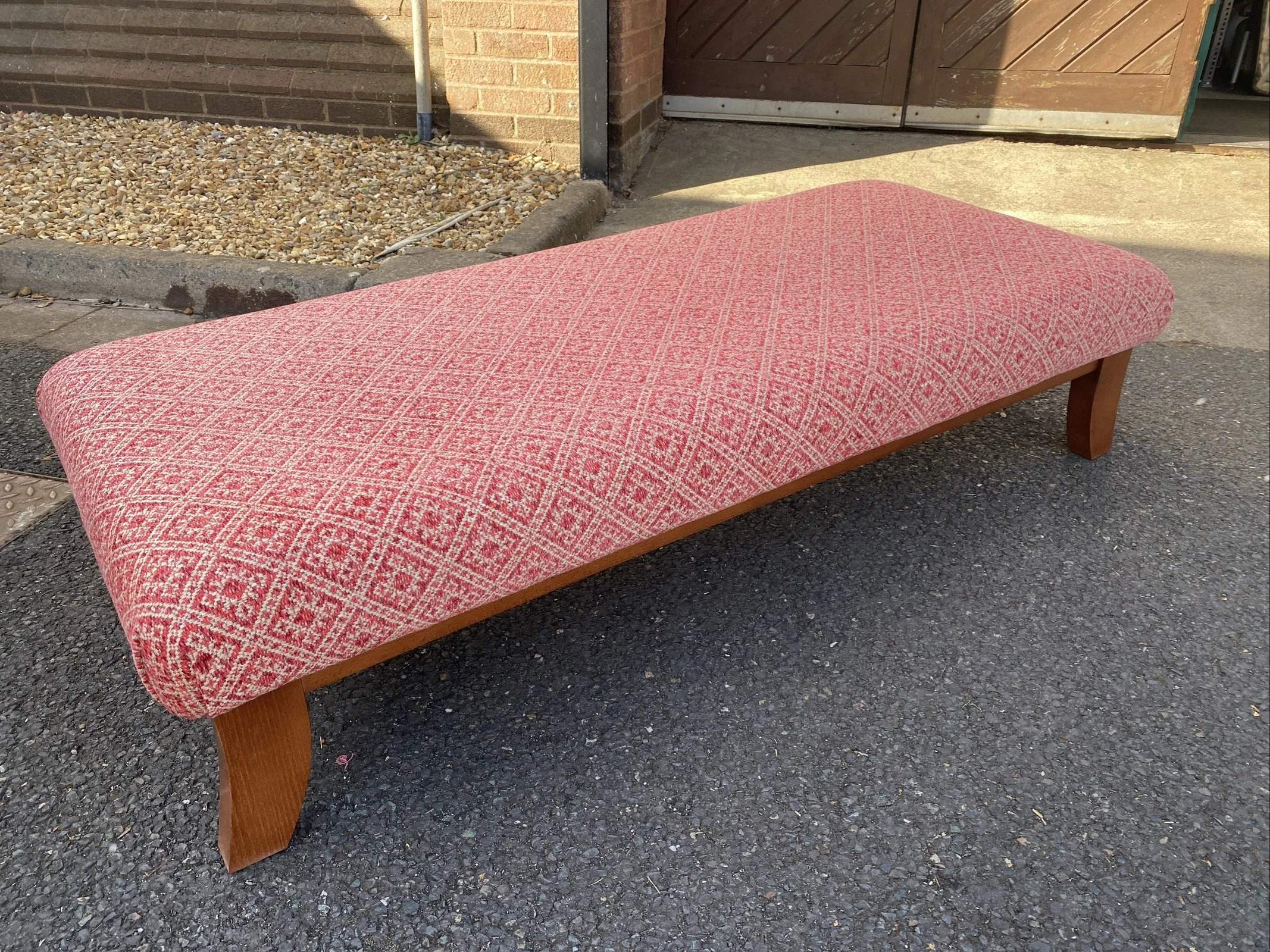 A pink patterned upholstered bench with wooden legs outdoors on asphalt pavement.