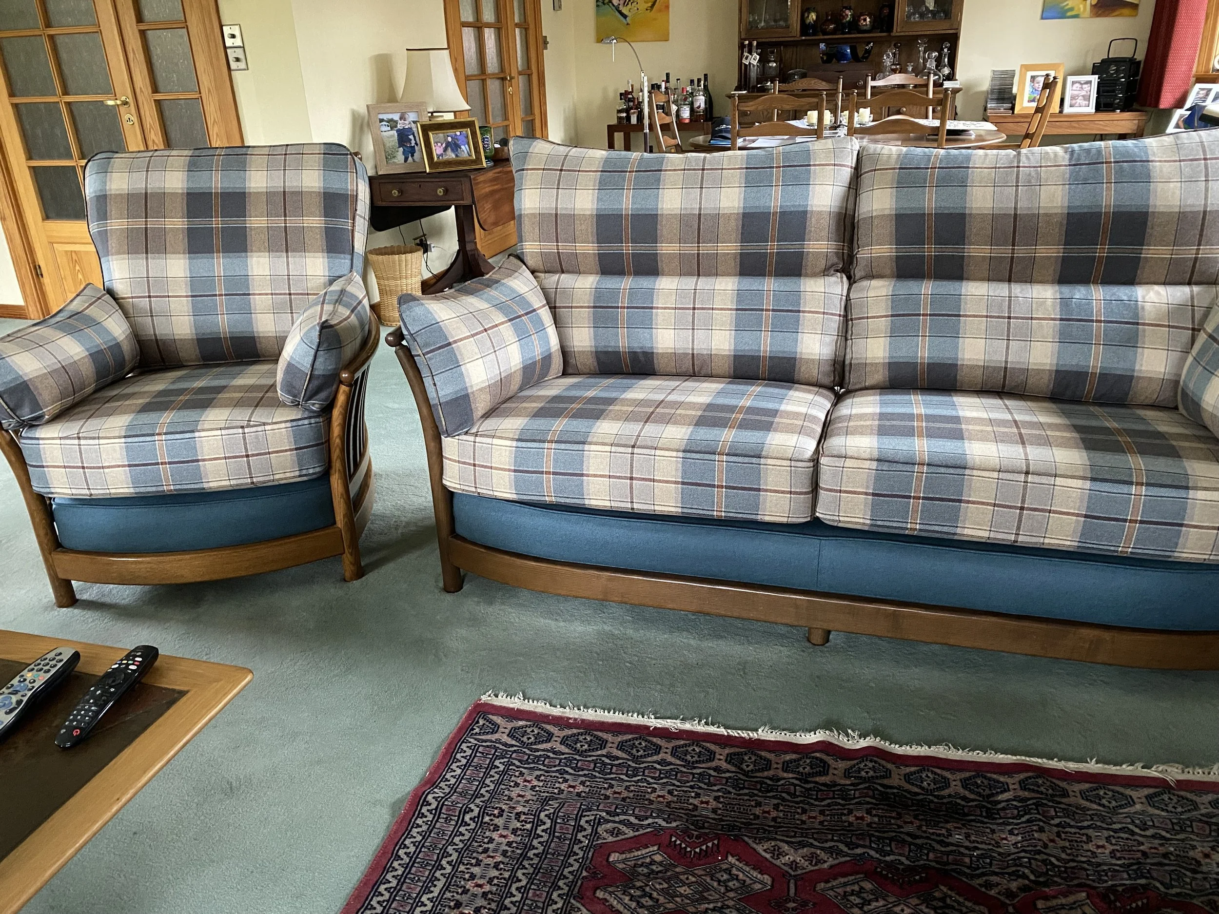 Living room with a plaid-patterned armchair and sofa, both with matching cushions, a wooden coffee table with remote controls, a carpet, and a background featuring a dining area with a wooden table and chairs, and a cabinet with bottles and picture f