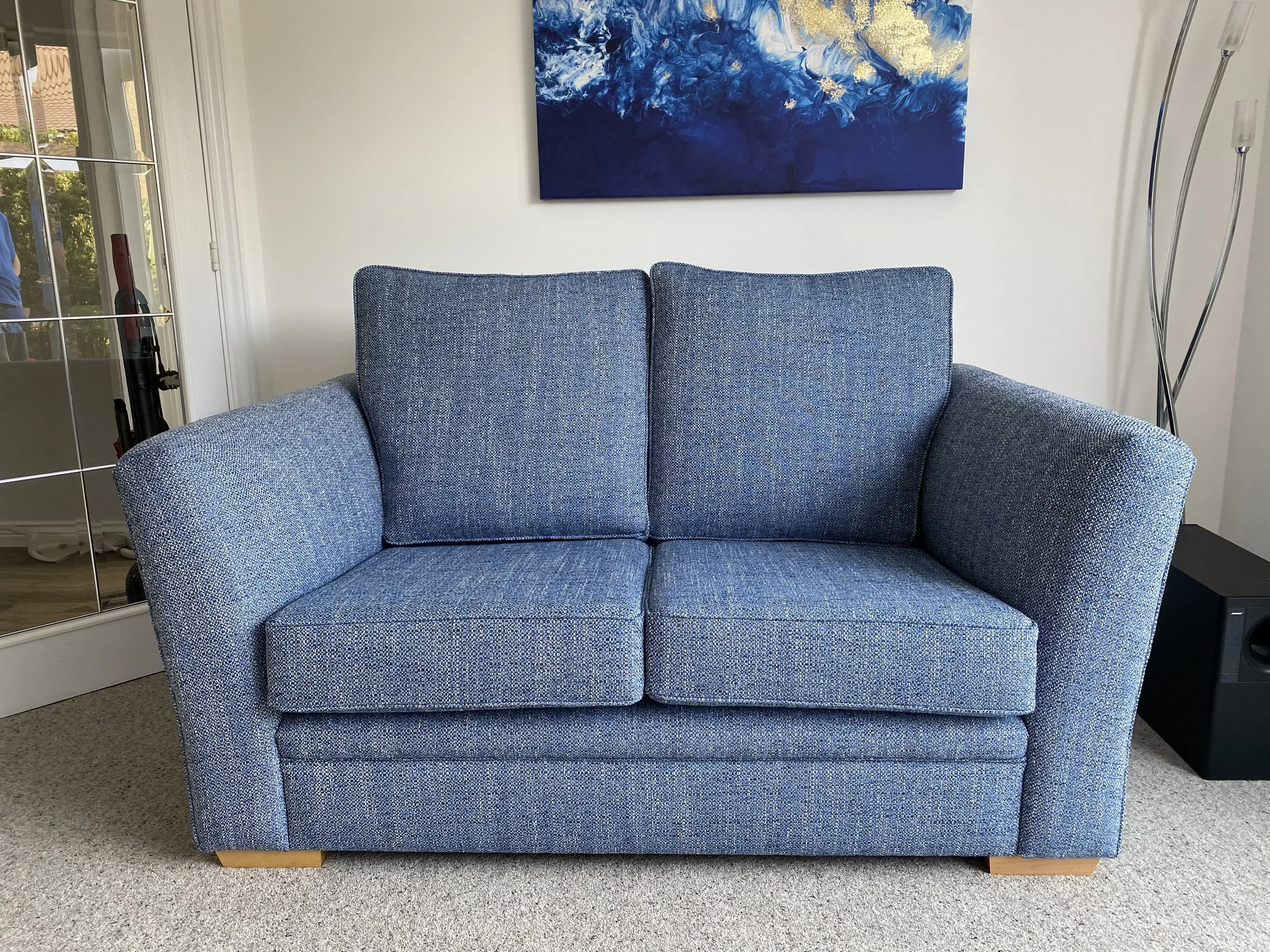 Blue upholstered two-seater sofa with wooden block legs in a living room, above which hangs a blue abstract painting. Part of a glass door and a tall lamp are visible beside the sofa.