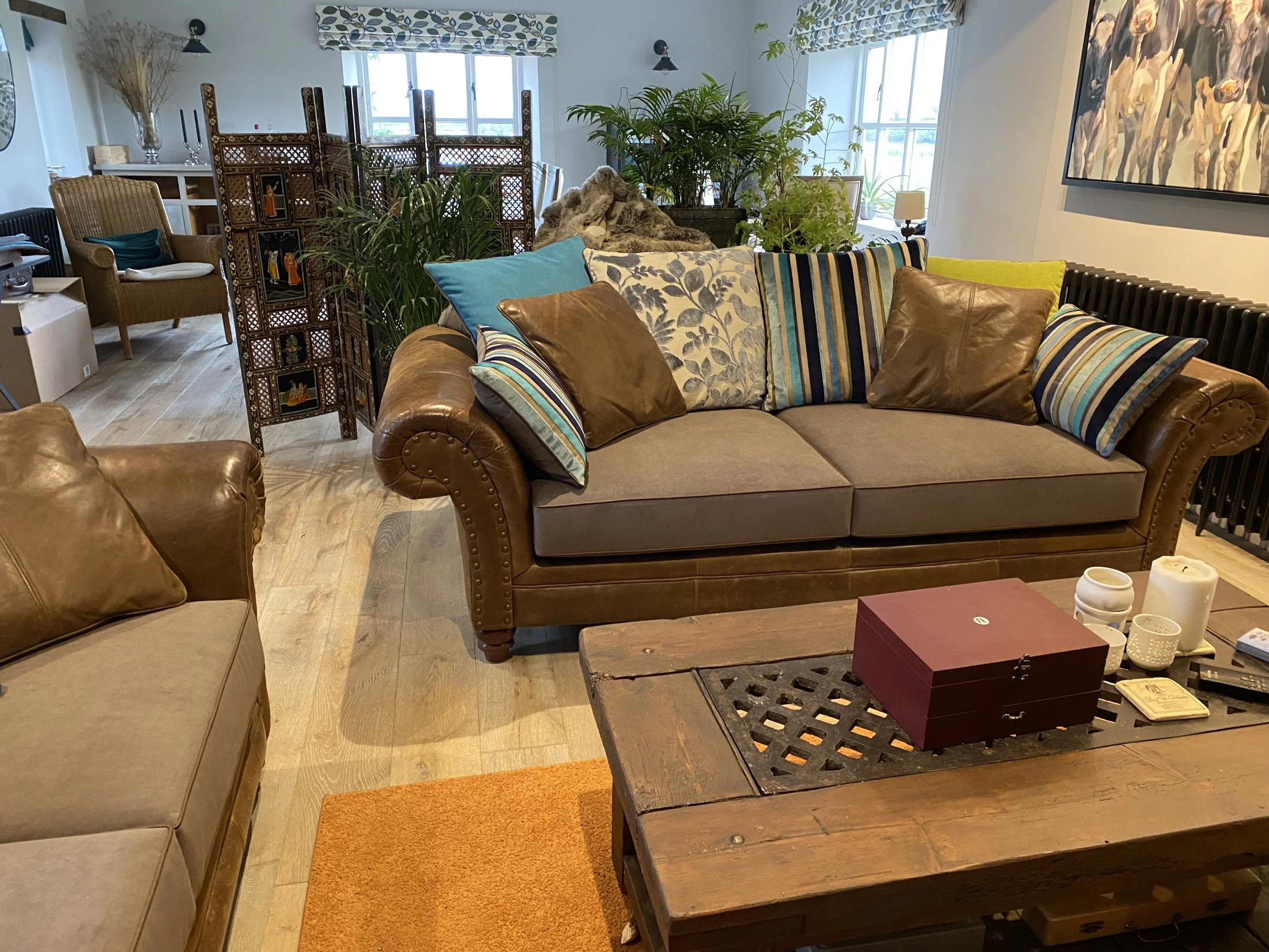 Living room with beige sofas, wooden coffee table, and various colorful cushions. There are windows with patterned curtains, plants, a decorative room divider, and artwork on the walls.