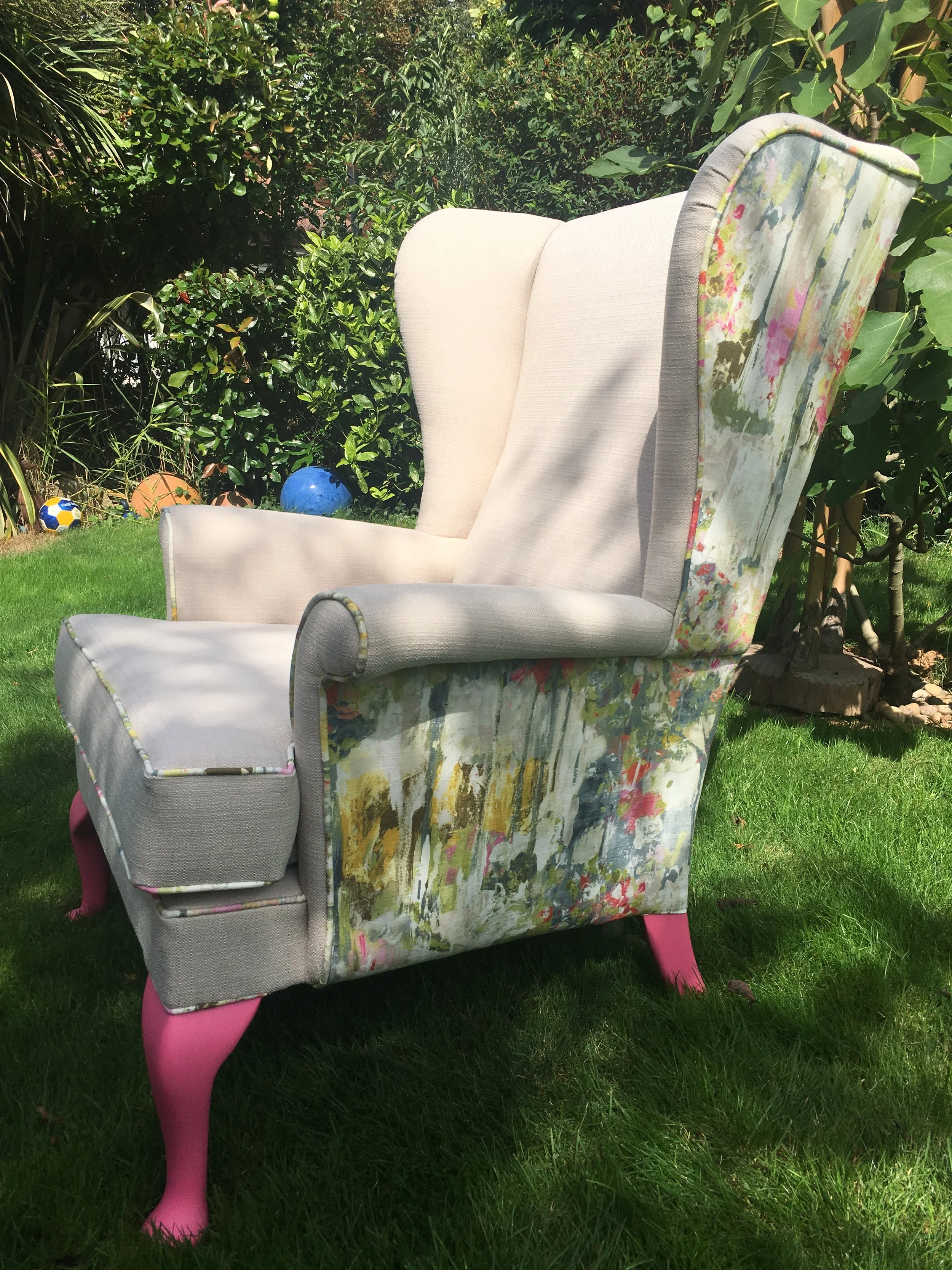 Colourful upholstered Parker Knoll 720 wingback armchair with pink legs on a grassy lawn with trees and bushes in the background.