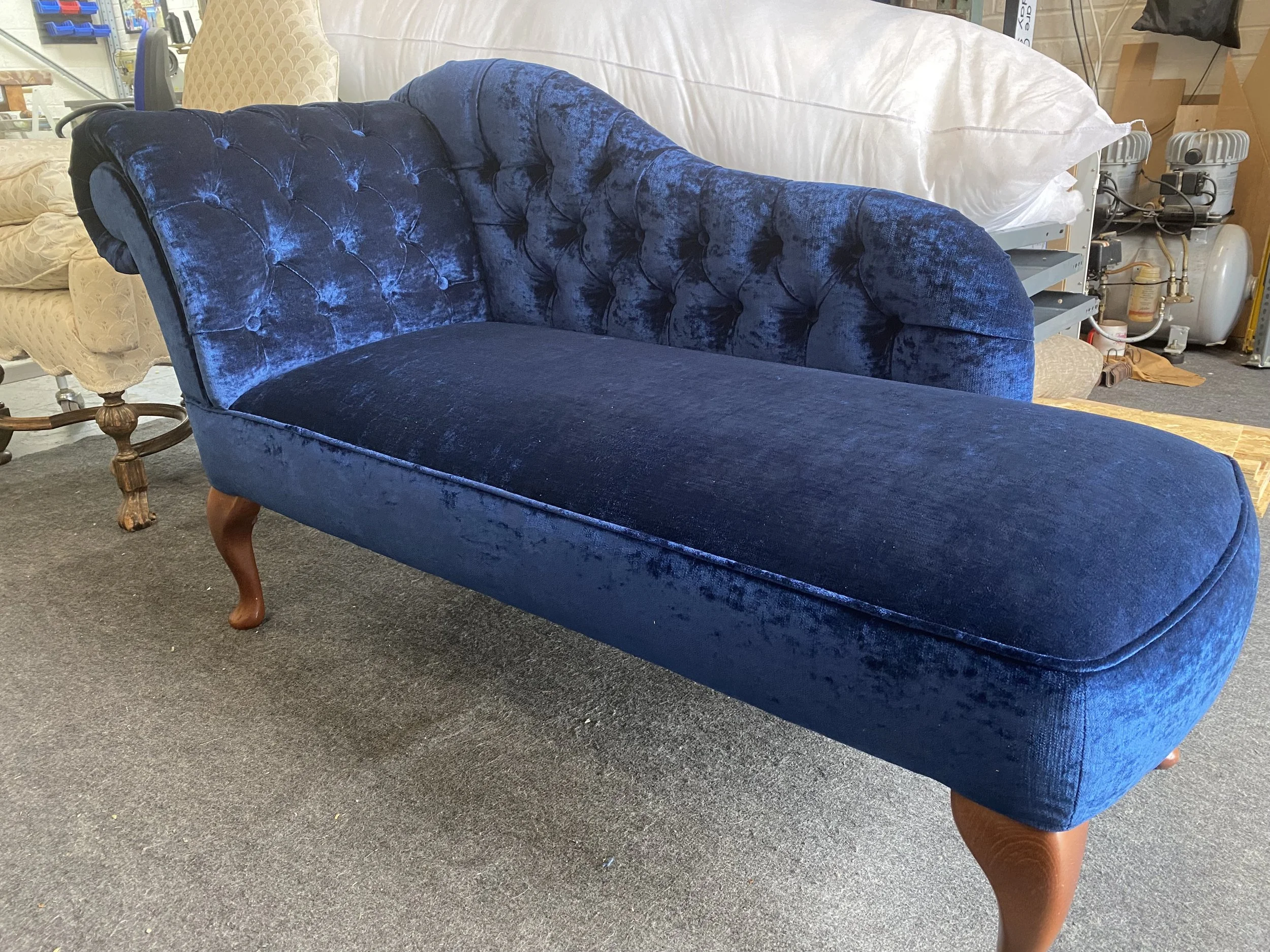 A vintage-style blue velvet chaise lounge with tufted backrest and curved wooden legs, situated in a furniture store.