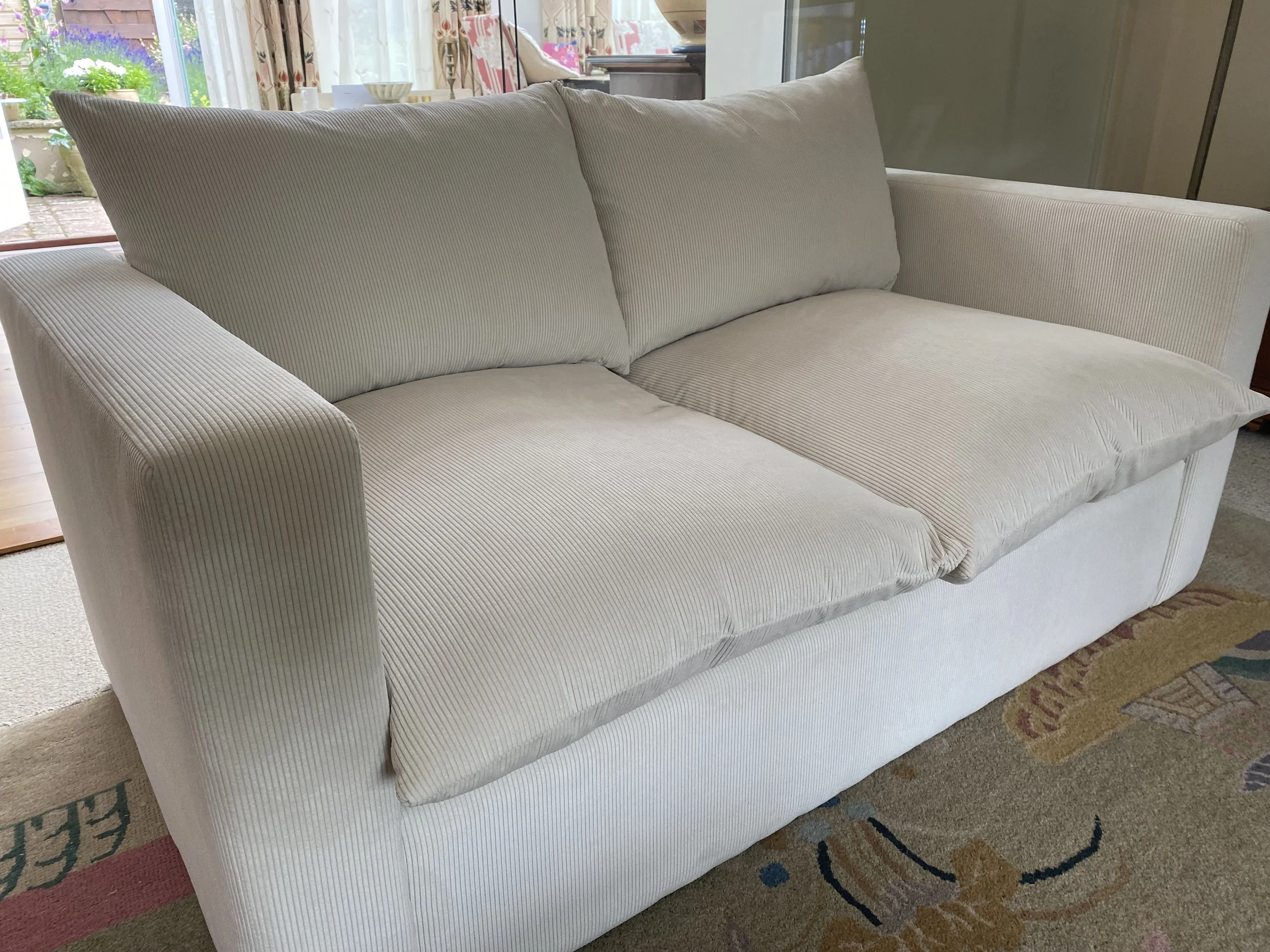 A cream-colored sofa with striped fabric in a living room, with a floral patterned rug underneath and a garden visible outside through glass doors in the background.