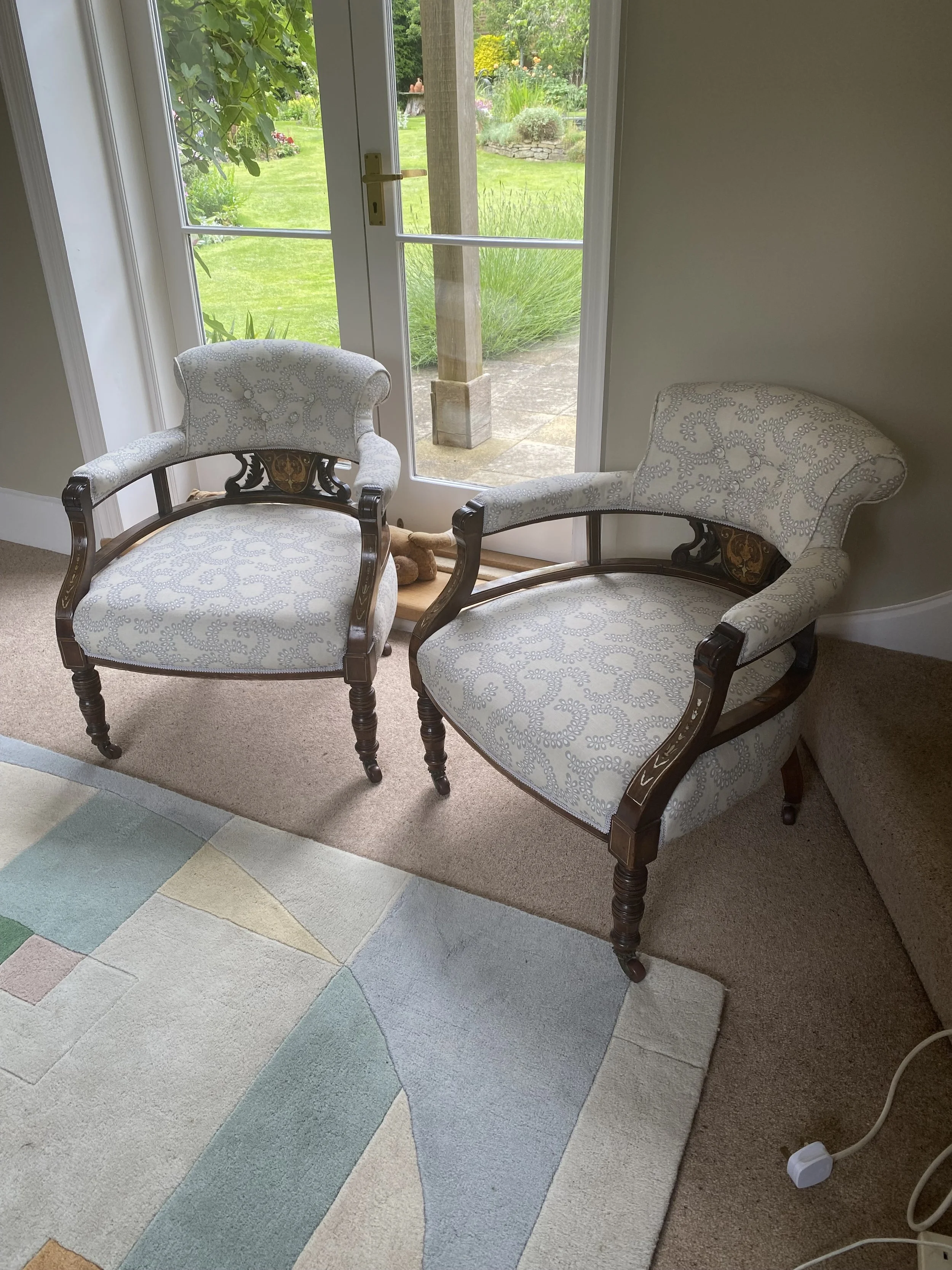 Two antique upholstered armchairs with wooden frames and patterned fabric, placed near a glass door leading to a garden with green grass and plants.