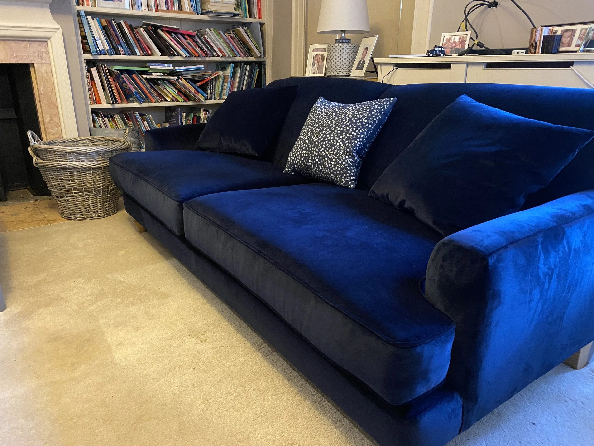 A dark blue velvet sofa with three cushions, one patterned and two plain, in a living room next to a white shelf with books and framed photos.