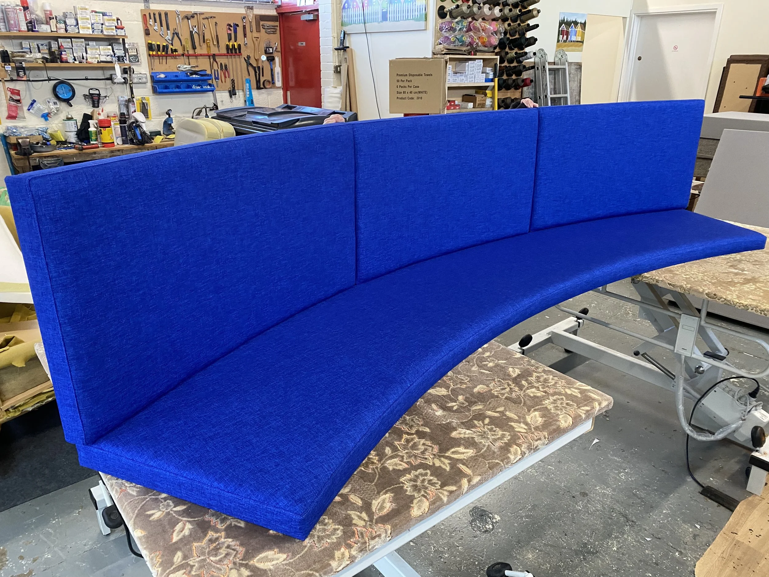 A vibrant blue upholstered bench with a curved seat and three backrest sections, positioned on a floral cushion atop a white massage or examination table in a workshop or upholstery studio environment.