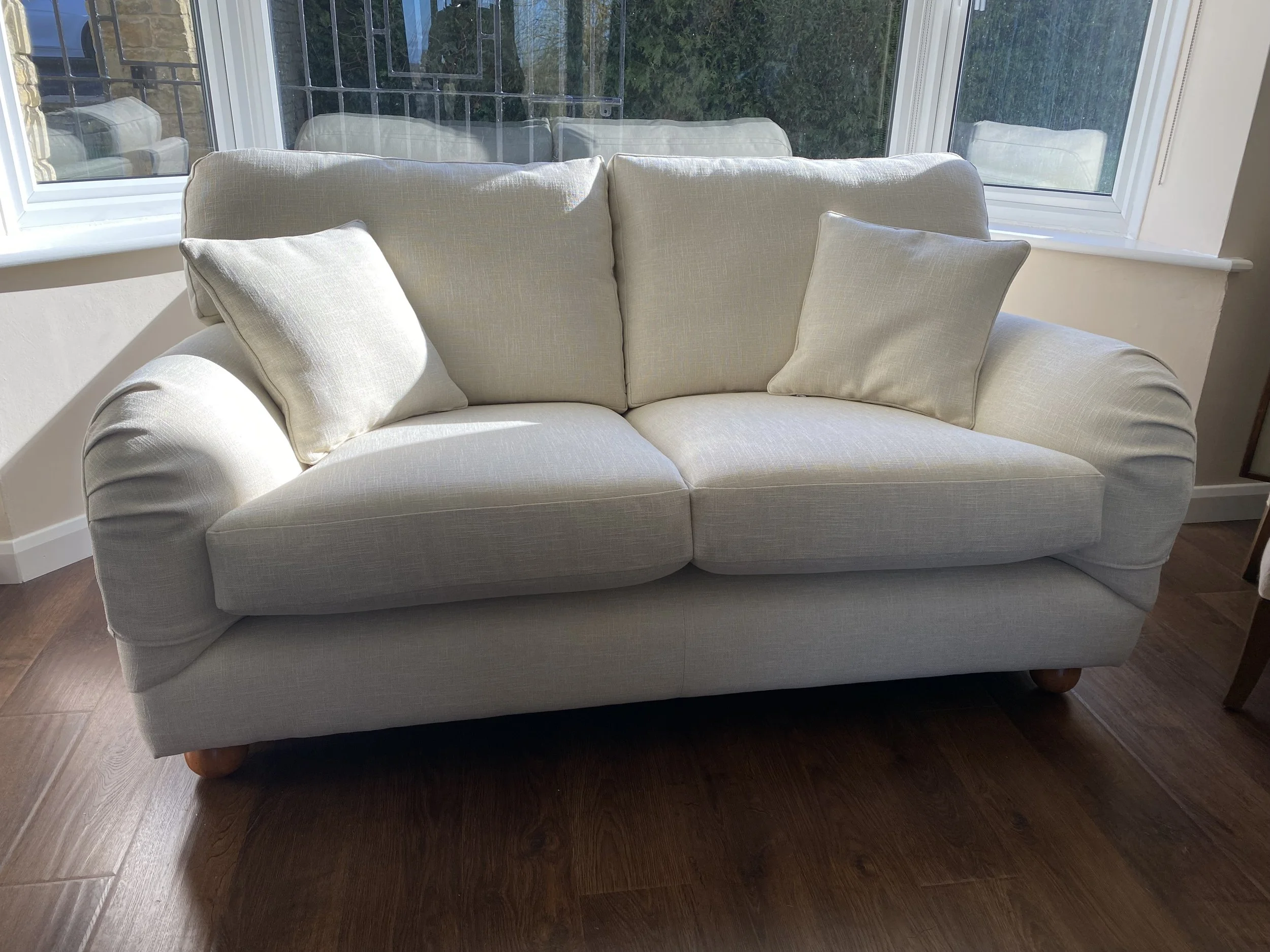 Cream-colored sofa with two matching throw pillows, positioned in front of large windows showing a green outdoor scene, on a dark wooden floor.