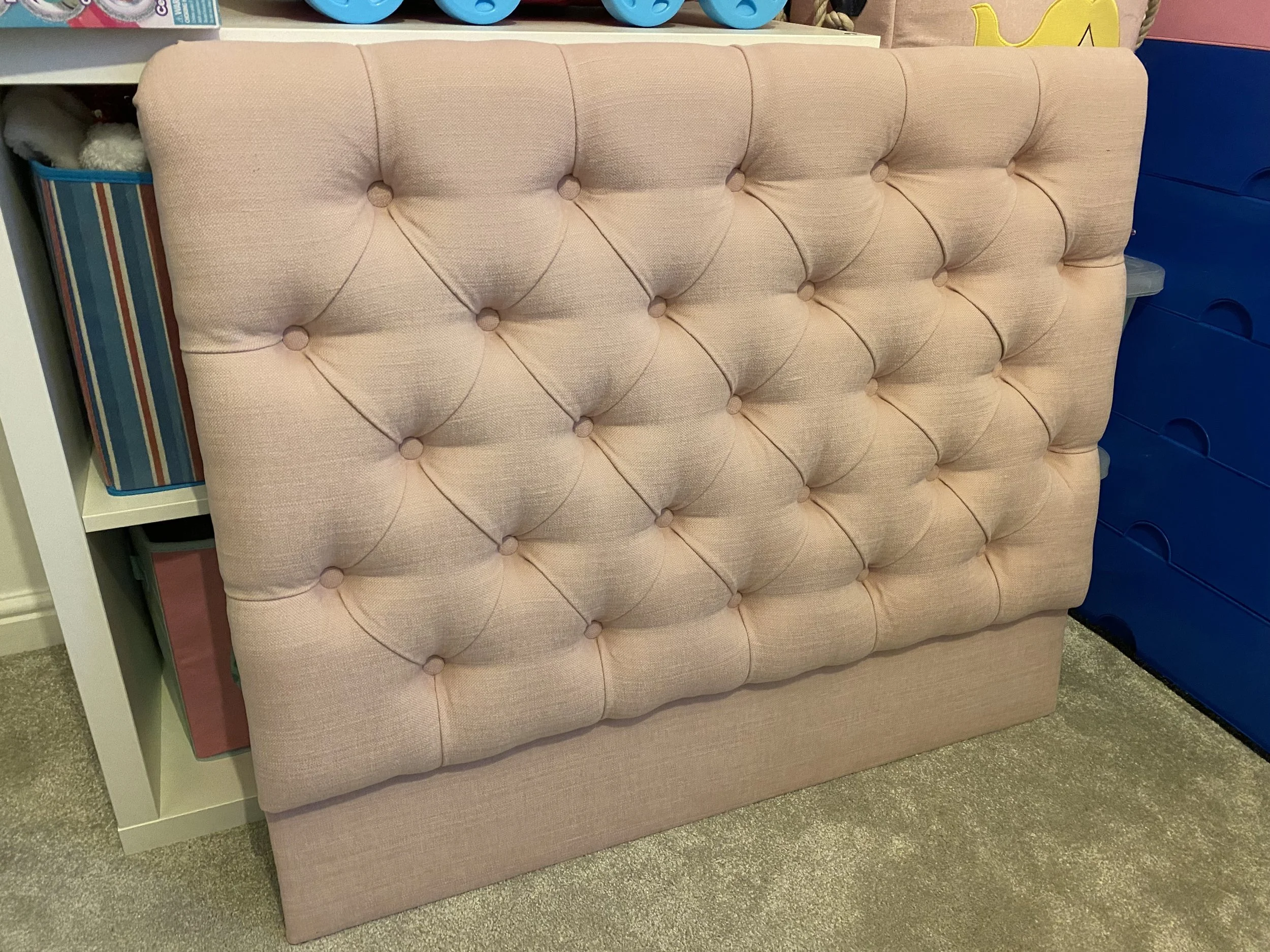 A pink upholstered tufted headboard with button details leaning against a shelf with storage bins.