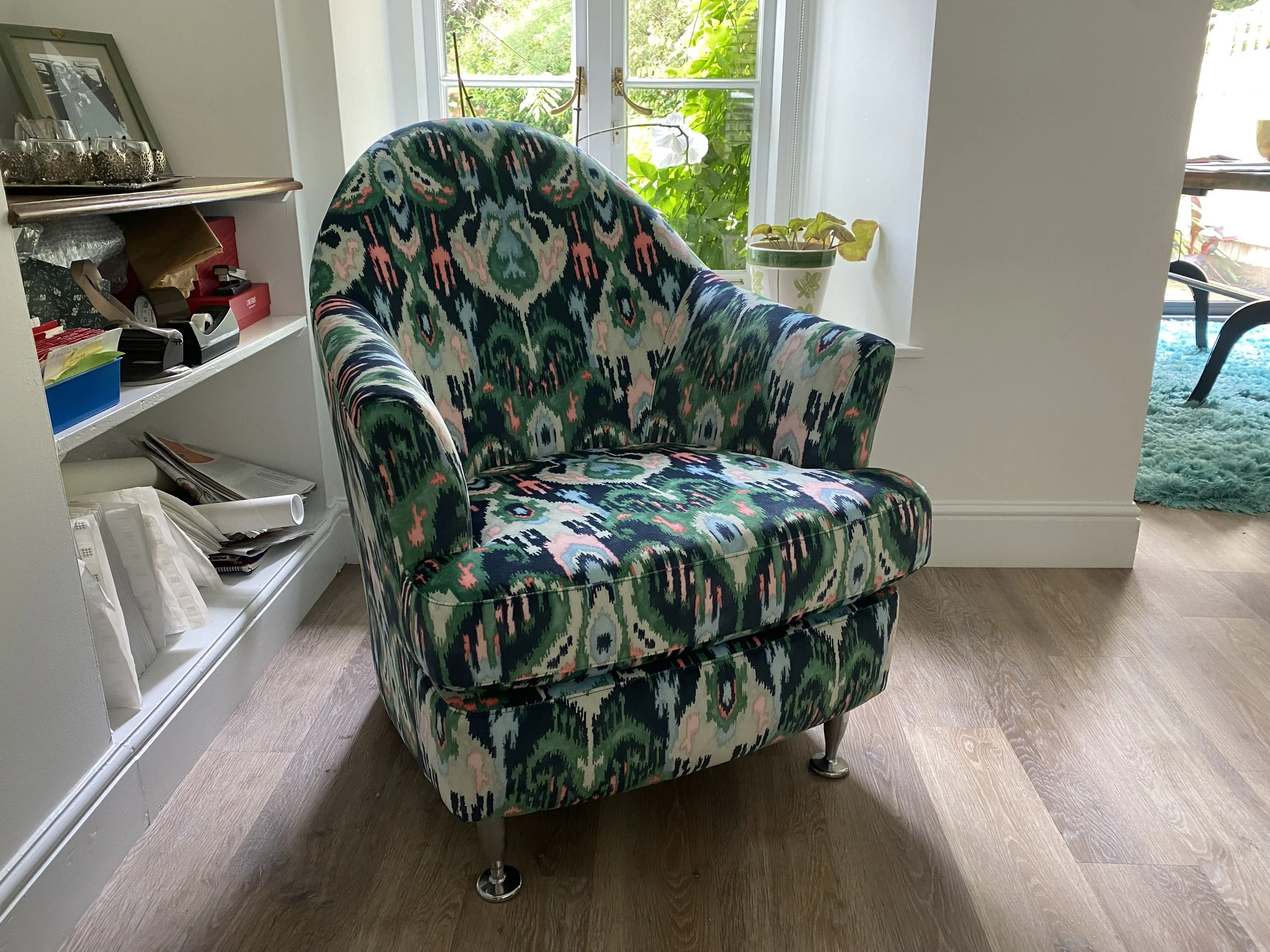 A colorful upholstered armchair with a patterned fabric in shades of green, pink, blue, and white, placed on a wooden floor near a window with natural light.