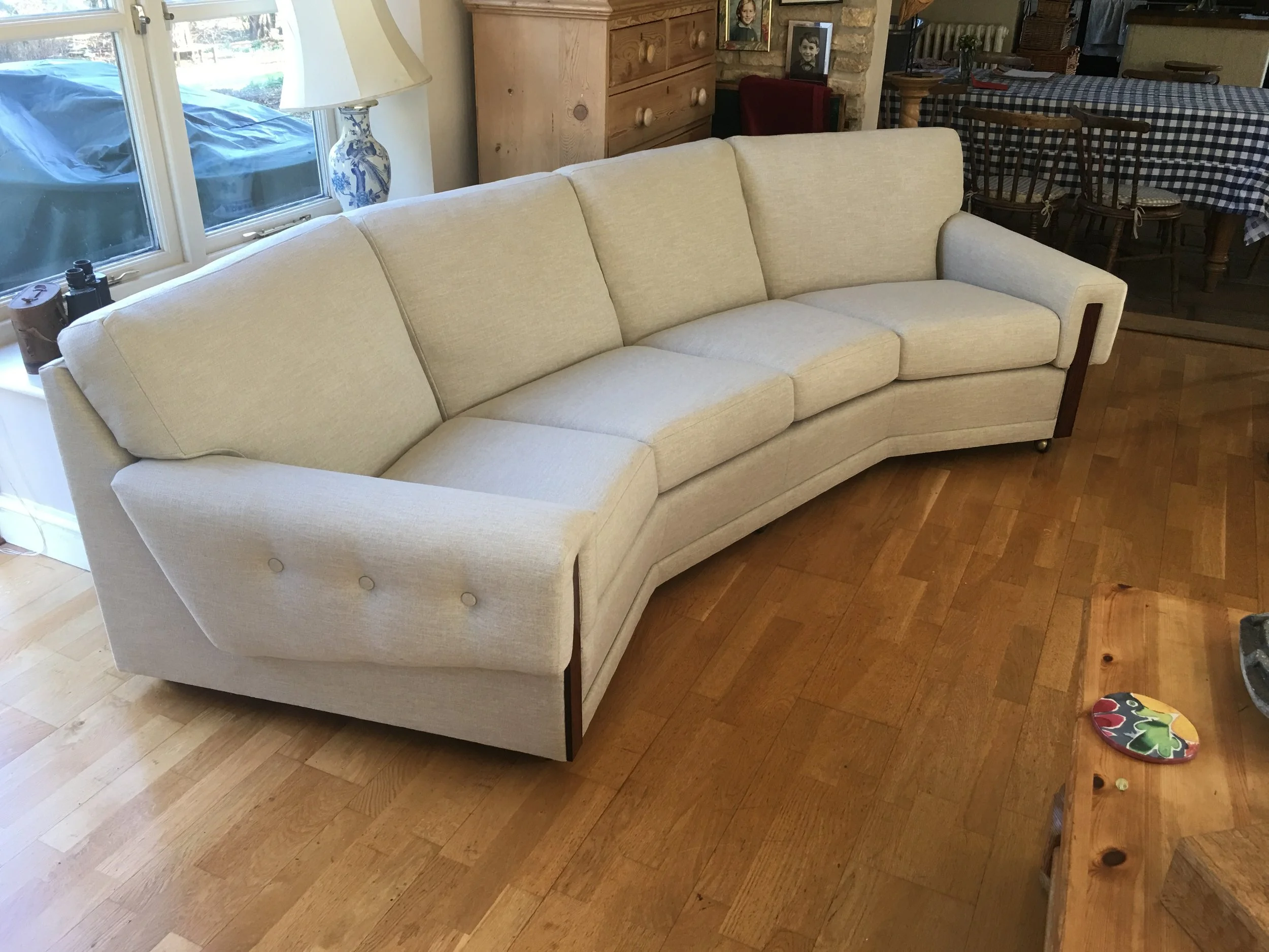 A beige vintage curved sofa in a living room next to a window with a white lampshade on a table nearby, and a wooden coffee table with a decorative plate and a small item on it, with a dining table and chairs in the background.