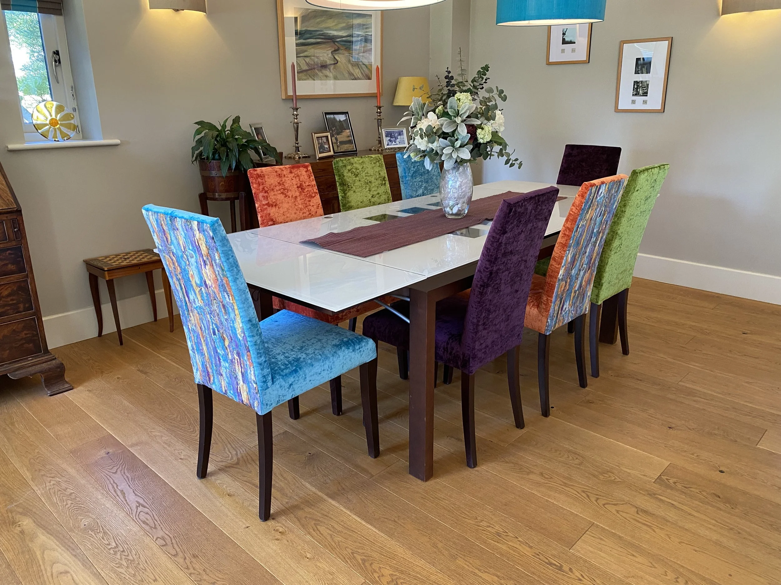 A dining room table with twelve colorful velvet chairs, a flower vase, and wall art.