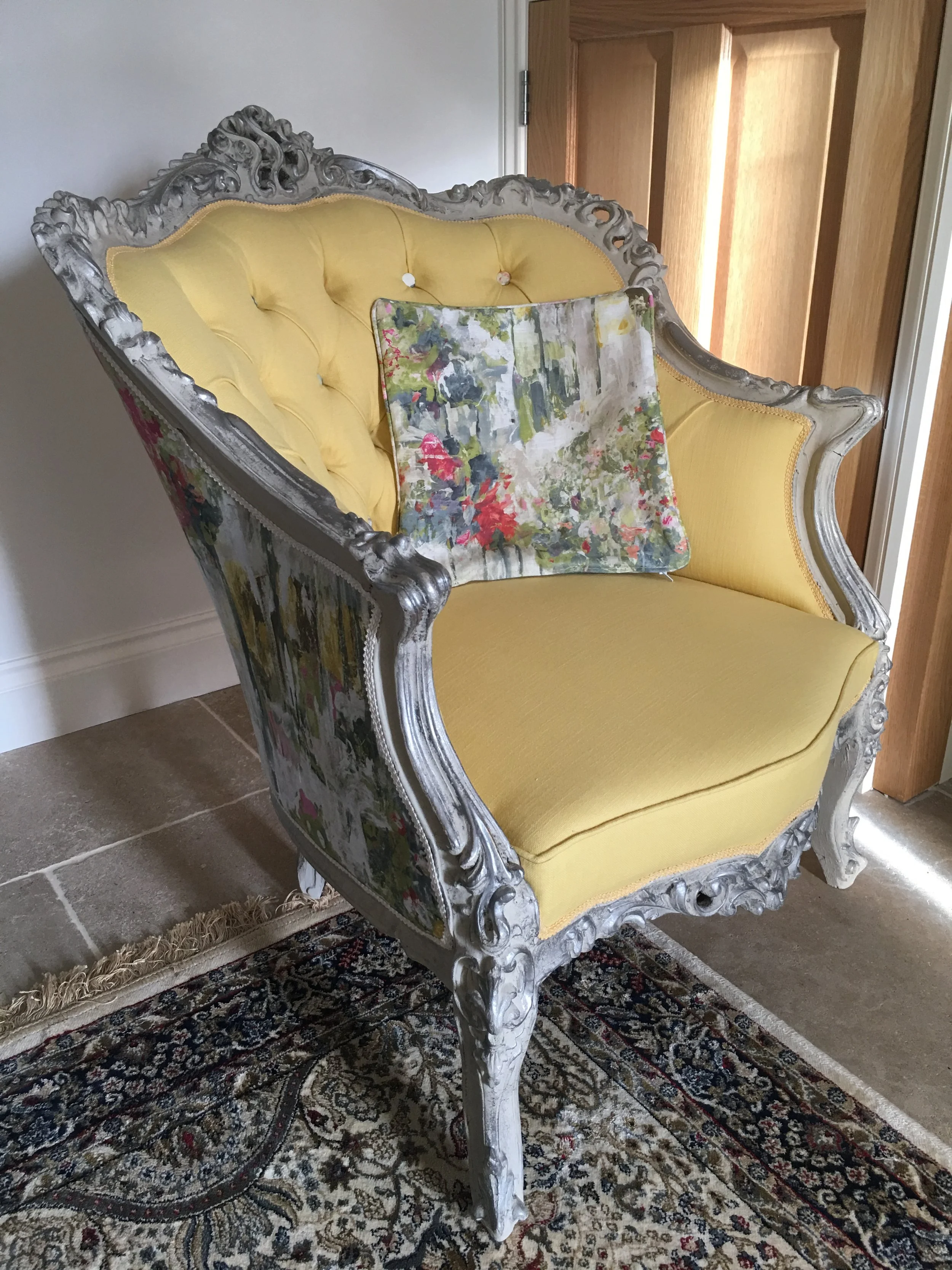 A vintage armchair with a yellow upholstered seat and back, ornate silver frame, and a small floral cushion, sitting on a patterned rug in front of a wooden door.