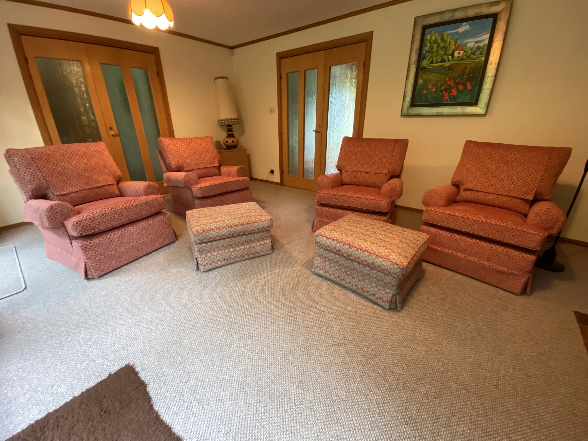 Living room with four pink patterned armchairs, two footstools, a wooden side table with a lamp, and a framed landscape painting on the wall.