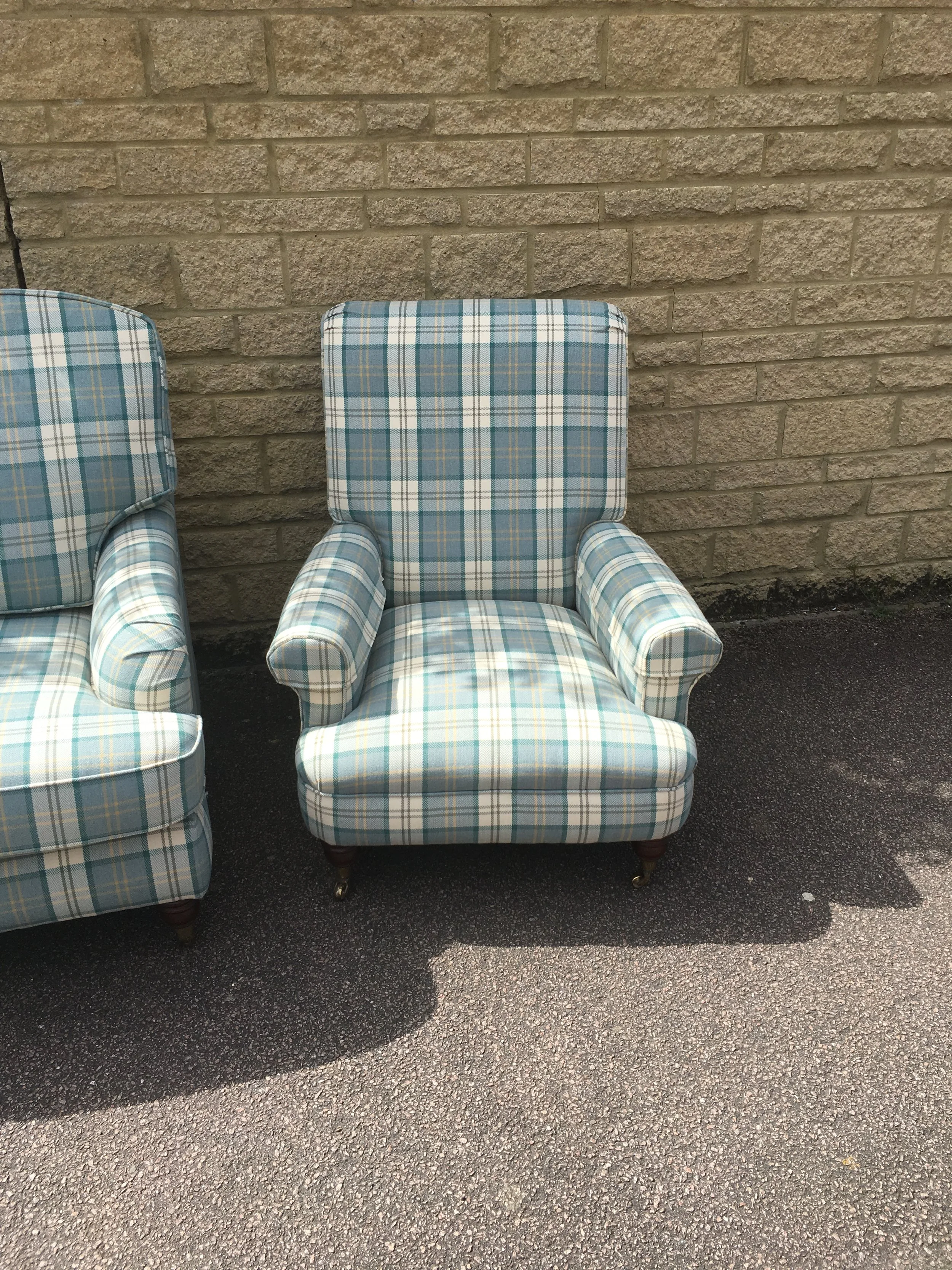 Patterned armchair with plaid fabric in shades of blue, green, and white, sitting outside on asphalt against a brick wall.