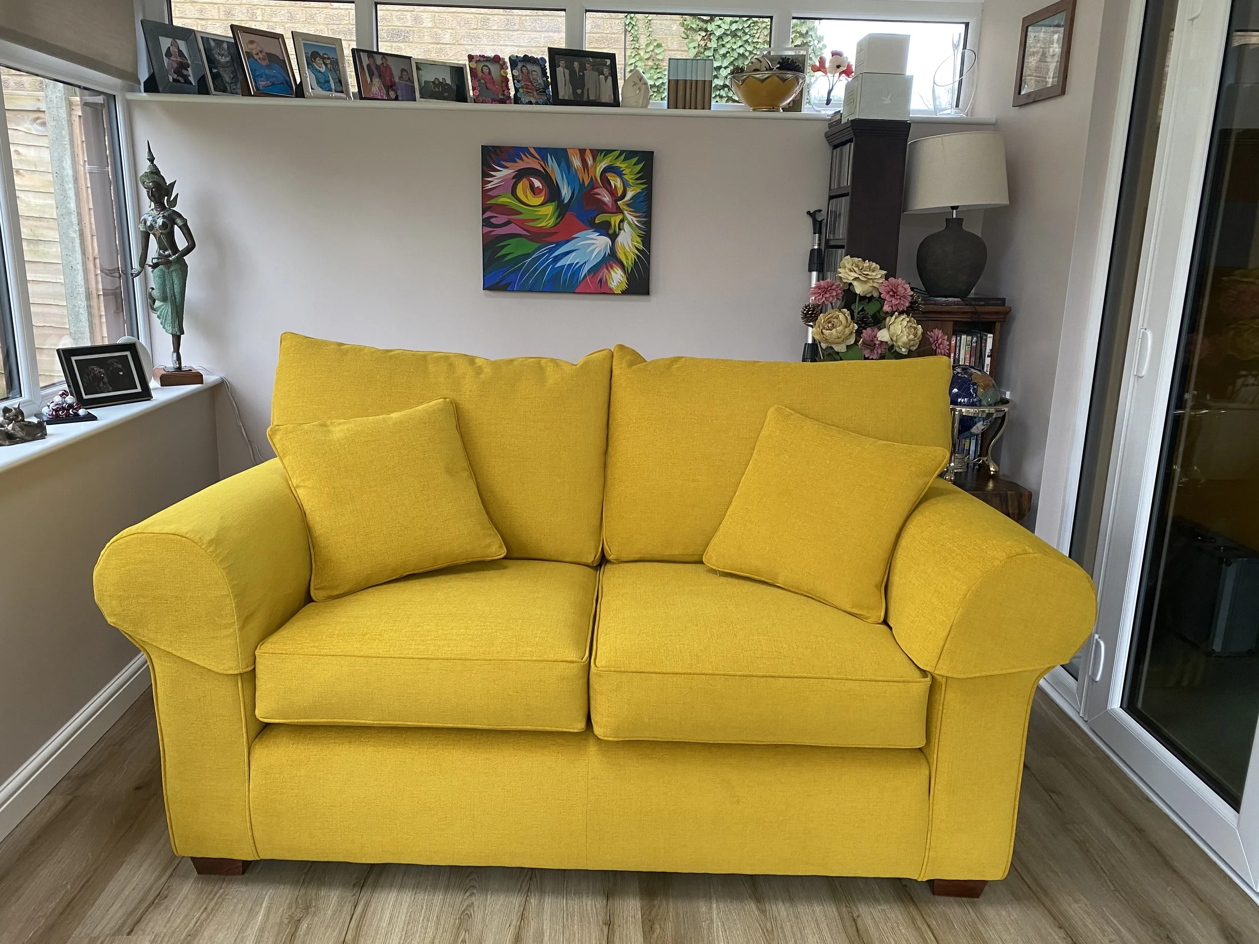 Yellow sofa with two matching cushions in a living room, with framed photos and a statue on the window ledge, a colorful lion portrait on the wall, and shelves with decorative items behind.