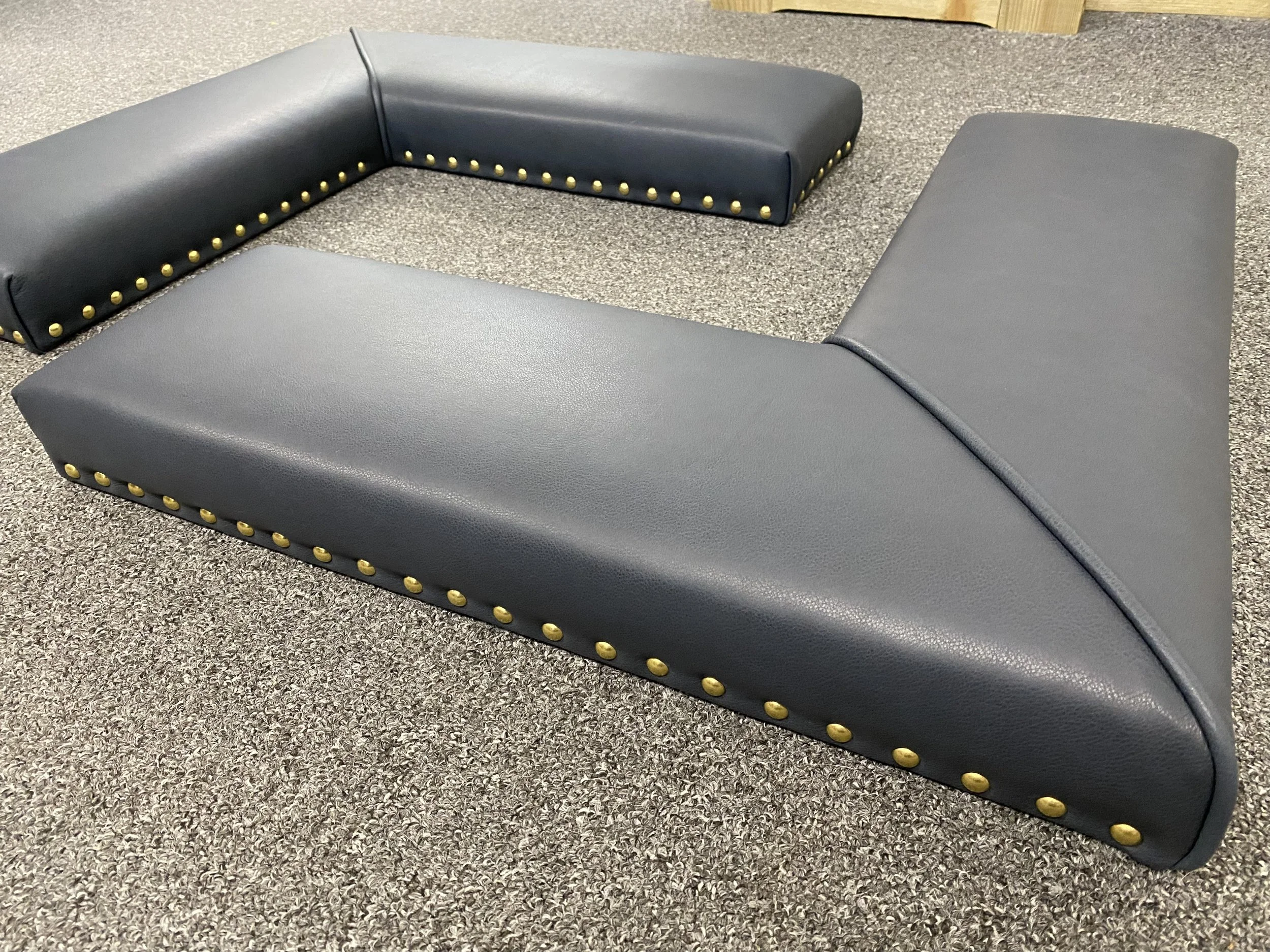 A set of foam padded, dark gray, L-shaped furniture pieces with gold nailhead trim, arranged on a carpeted floor.