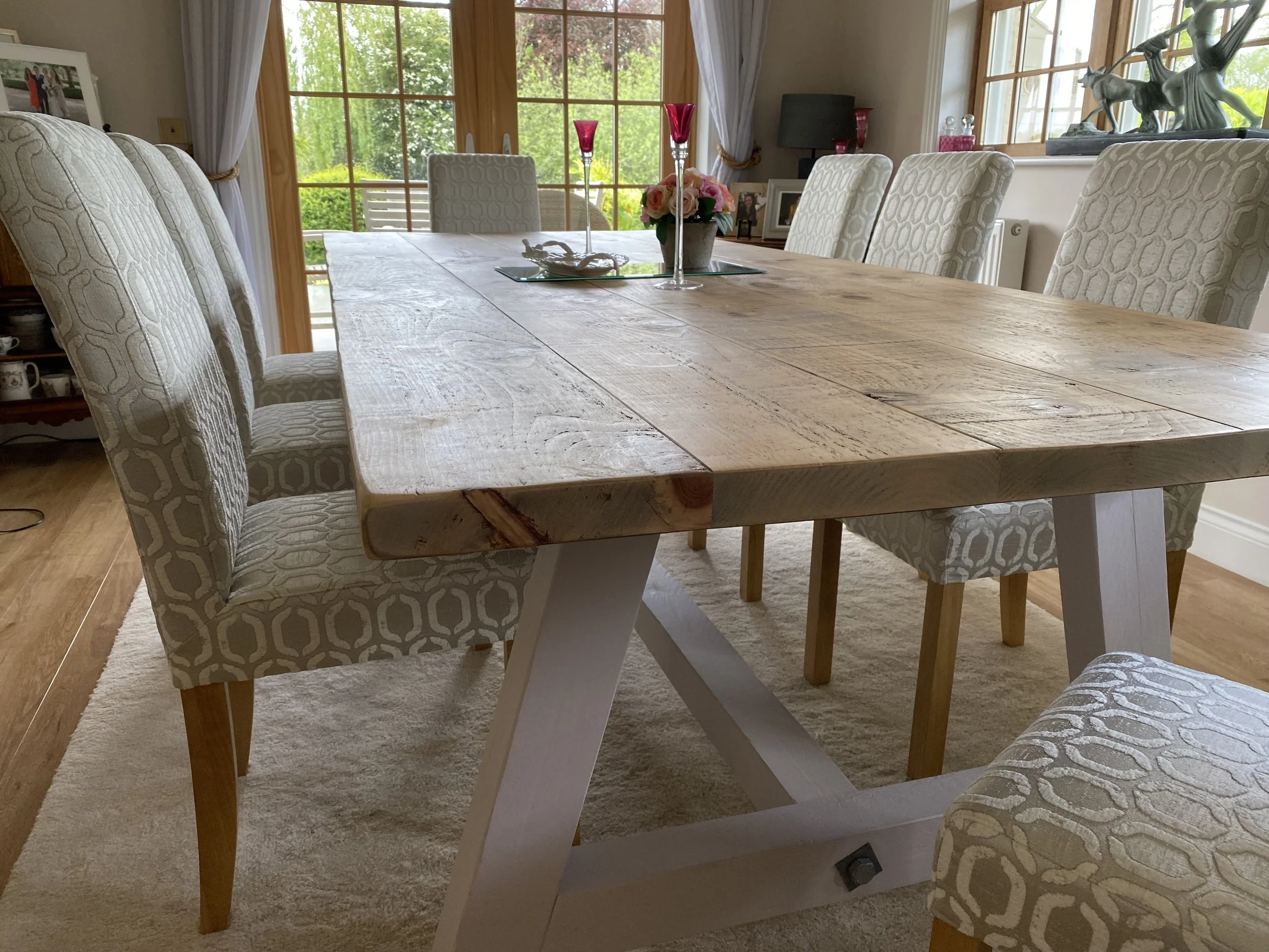 Wooden dining table with six upholstered chairs in a brightly lit room with large windows and a garden view.