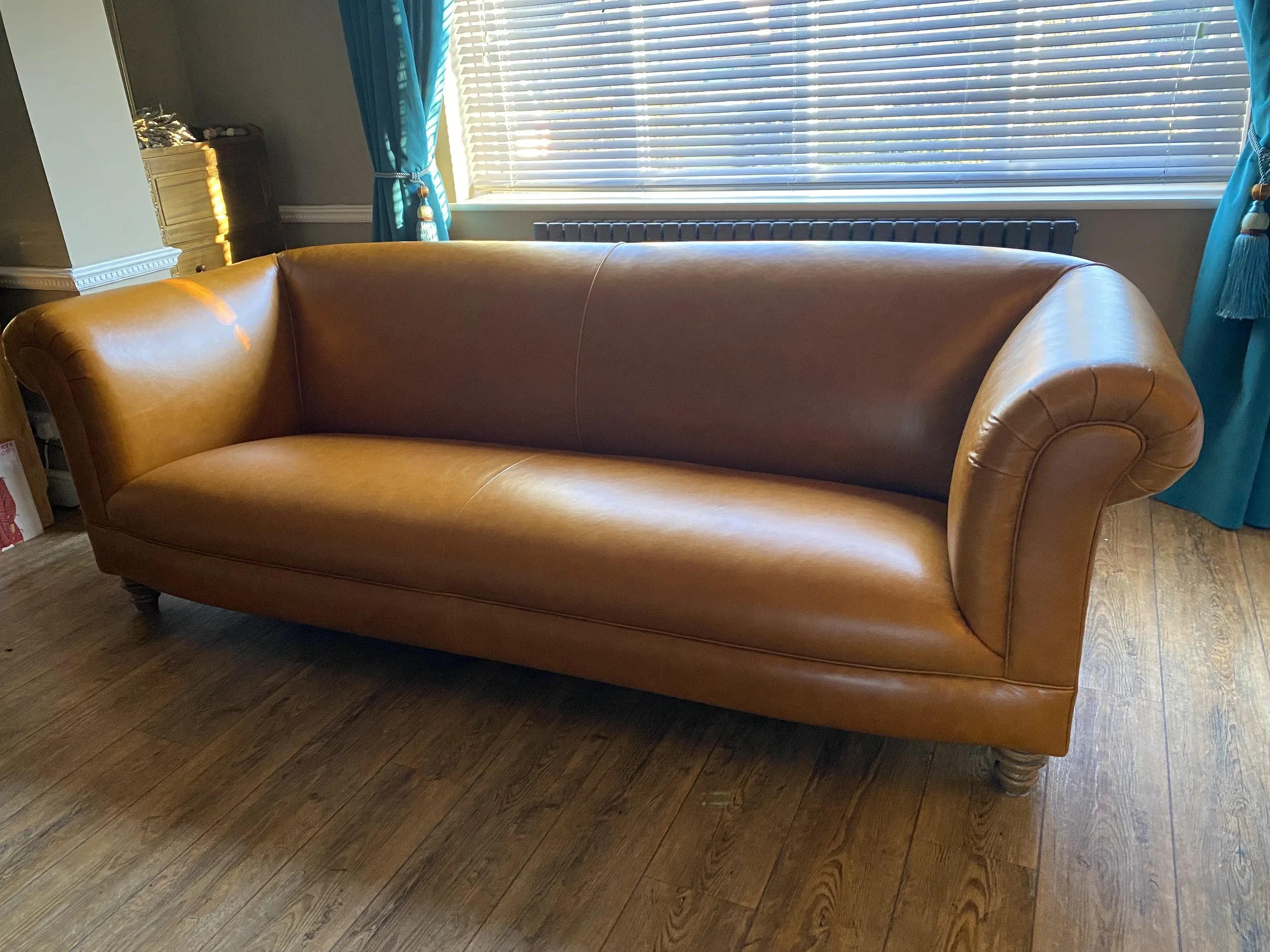 A brown leather sofa with rolled arms and wooden legs in front of a window with closed blinds and turquoise curtains.