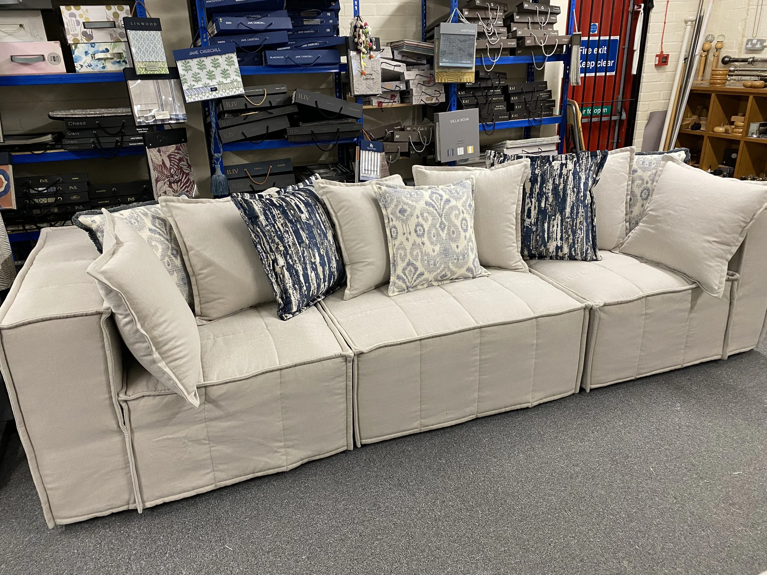 Beige sectional sofa with multiple decorative pillows in shades of cream, white, and blue, placed in a furniture store with shelves stacked with boxes and other furniture items in the background.