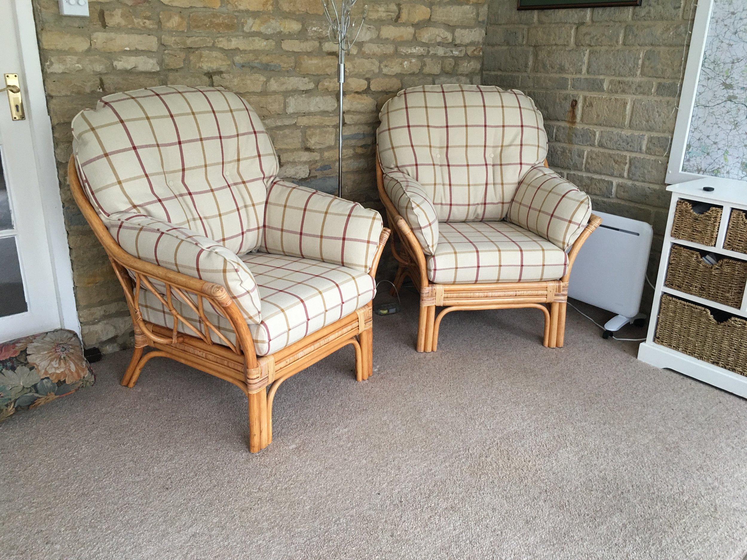 Two rattan armchairs with plaid cushions on a beige carpet in a room with a brick wall. There is a white shelving unit with woven baskets and a small heater in the background. A floor lamp is between the chairs.