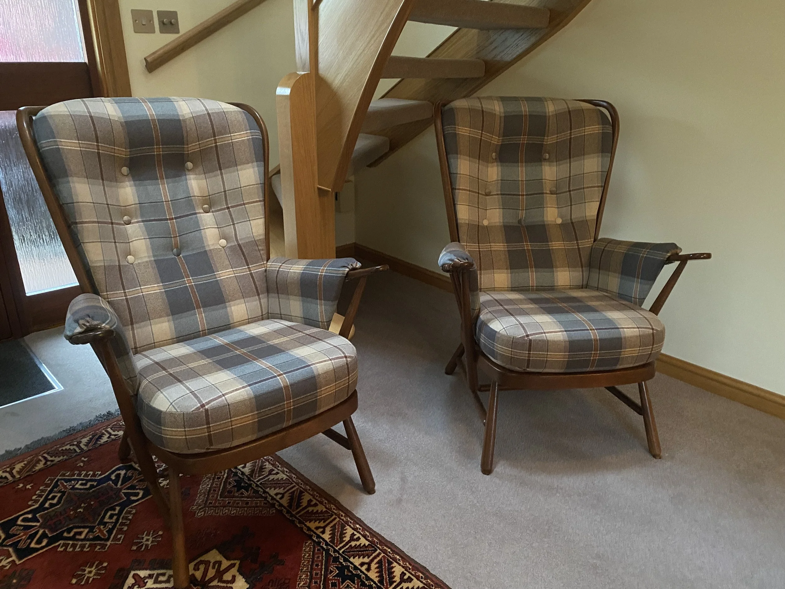 Two vintage plaid armchairs with wooden frames and armrests, positioned on a beige carpet near a small rug with a geometric pattern, in a room with light walls and a staircase.