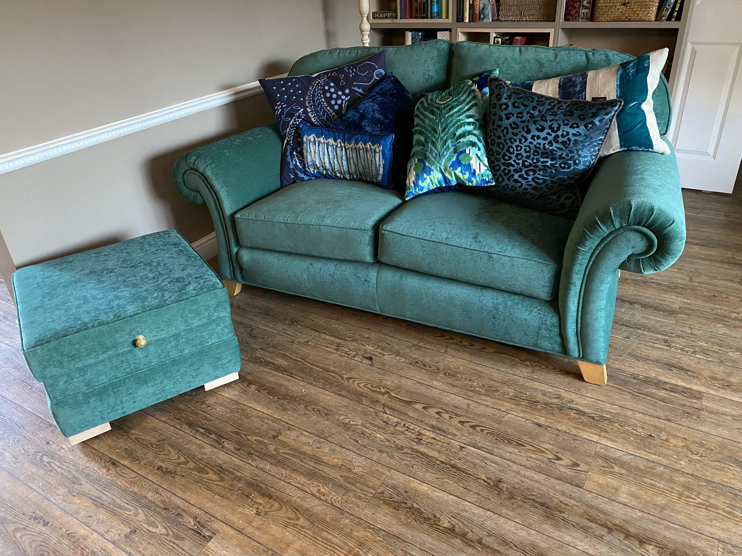 Green velvet sofa with multiple colorful patterned throw pillows, accompanied by a matching green velvet ottoman on a wooden floor, with a bookshelf in the background.