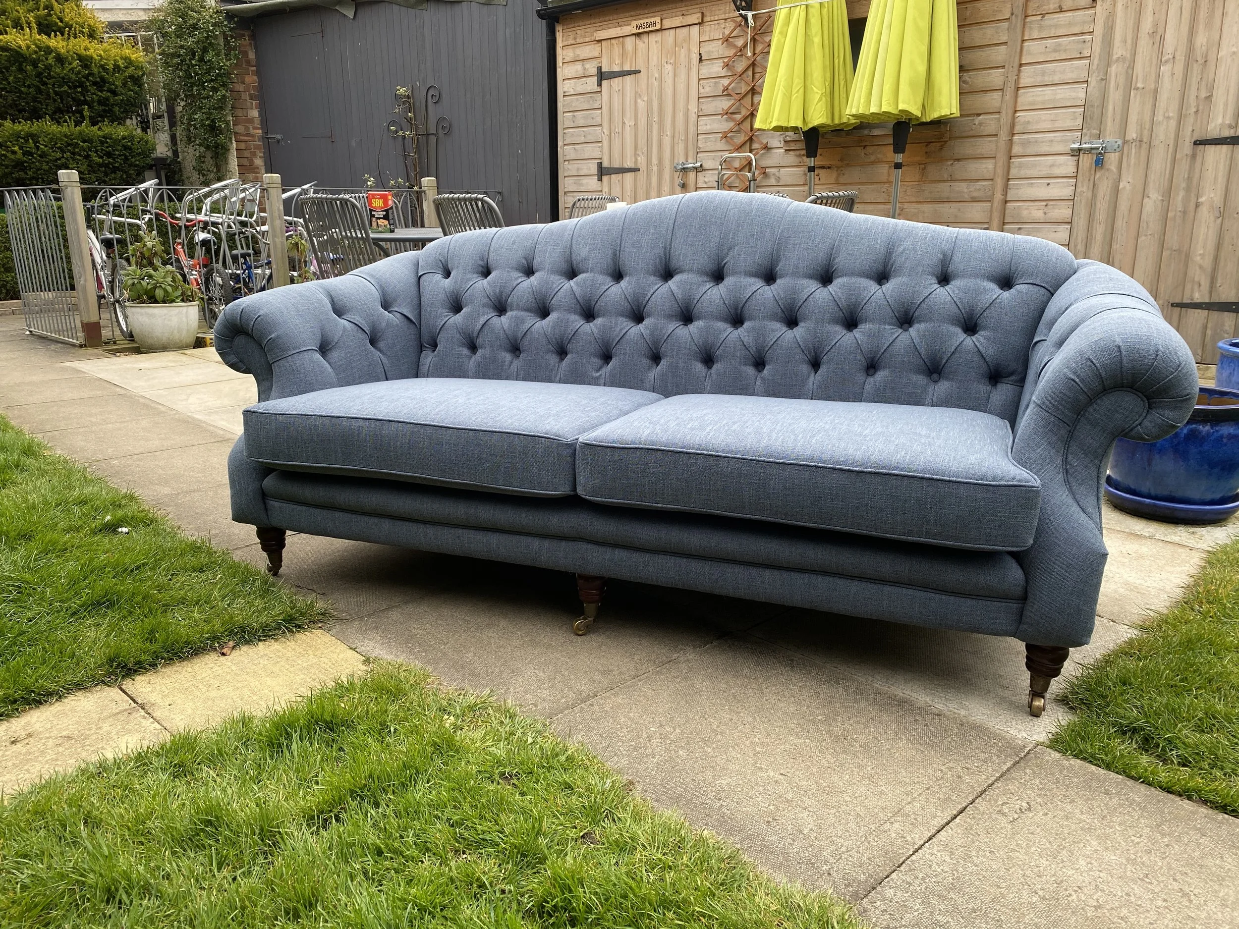 A vintage blue tufted sofa placed on a concrete pavement in a backyard with grass on both sides and a wooden fence in the background.