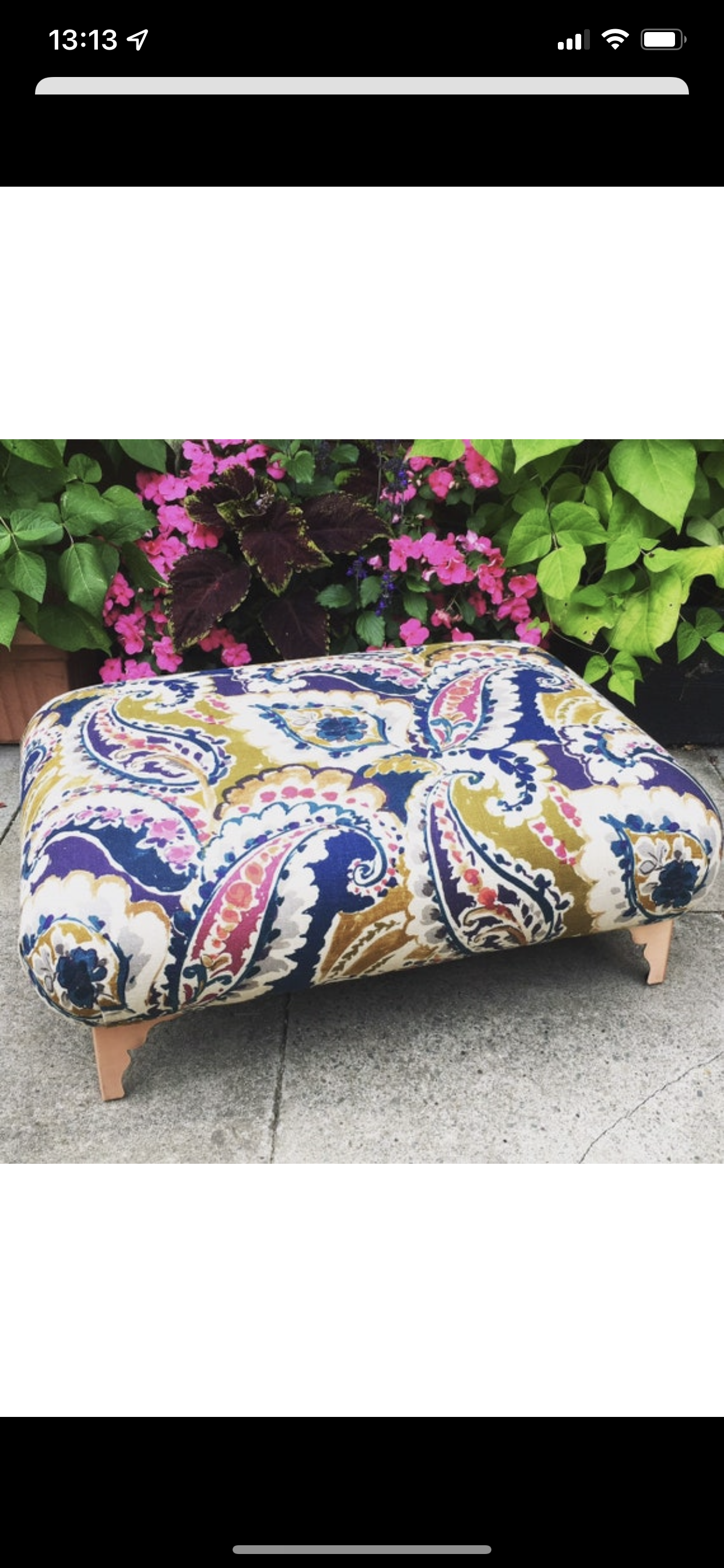 A colorful upholstered ottoman with a floral and paisley pattern, placed on a concrete surface, with pink and green leaves and flowers in the background.
