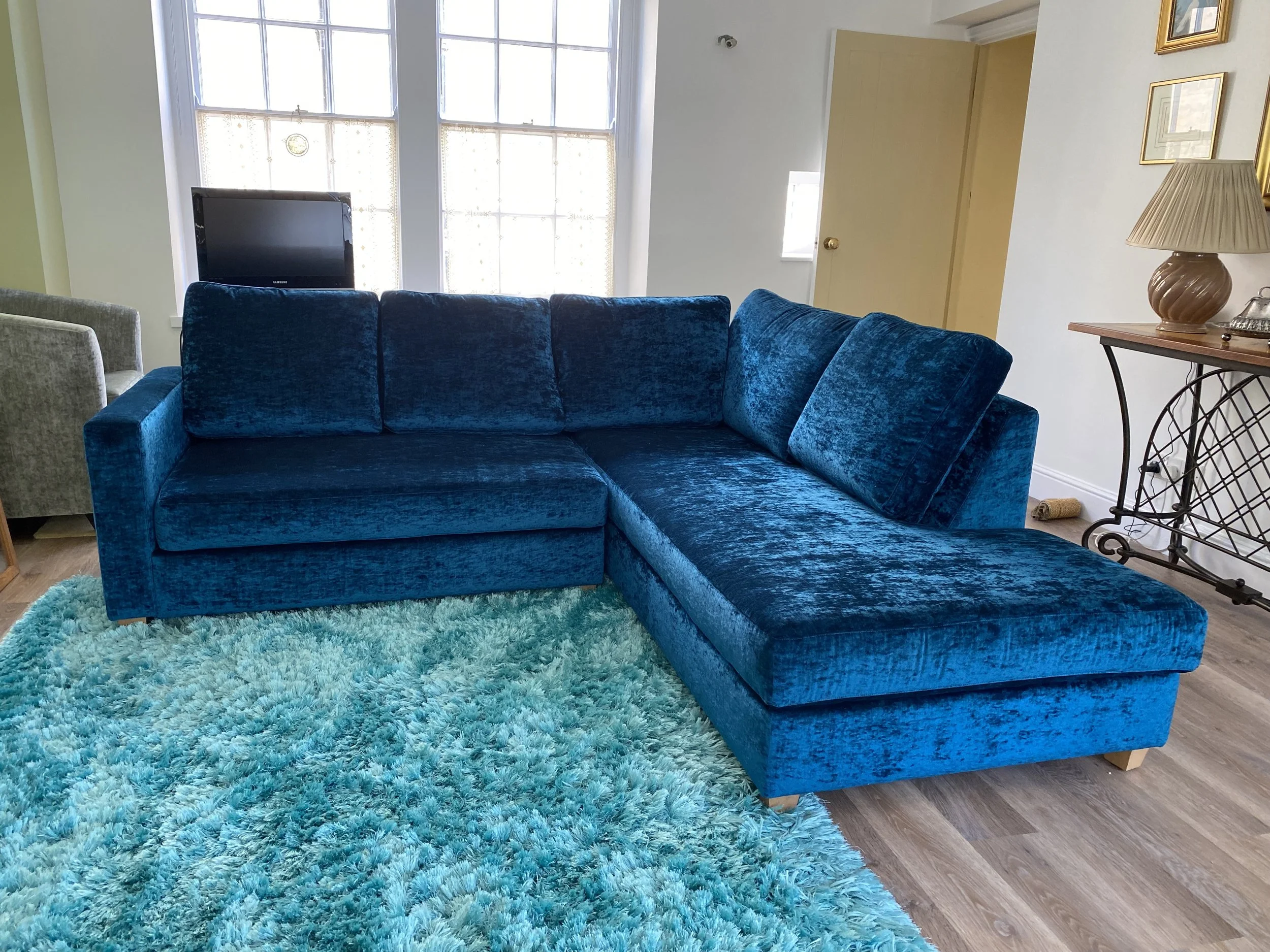 A bright living room with a large blue velvet sectional sofa, a turquoise shag rug, and a window with curtains behind the sofa.