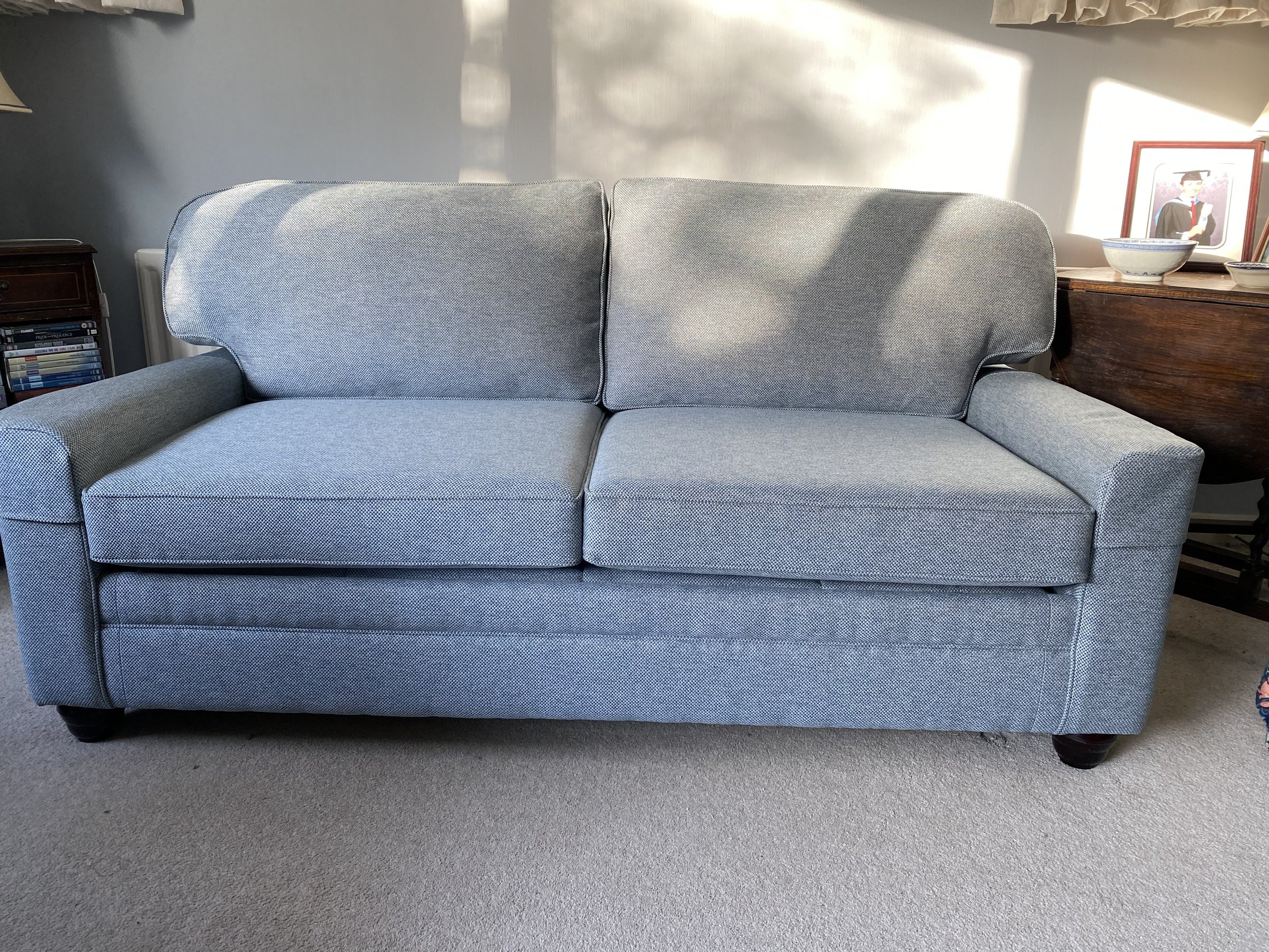 Light blue fabric sofa with two seat cushions and two back cushions, wooden legs, in a living room with a beige carpet. There is sunlight on the wall behind the sofa. To the right, there is a wooden side table with a framed picture of a graduation in