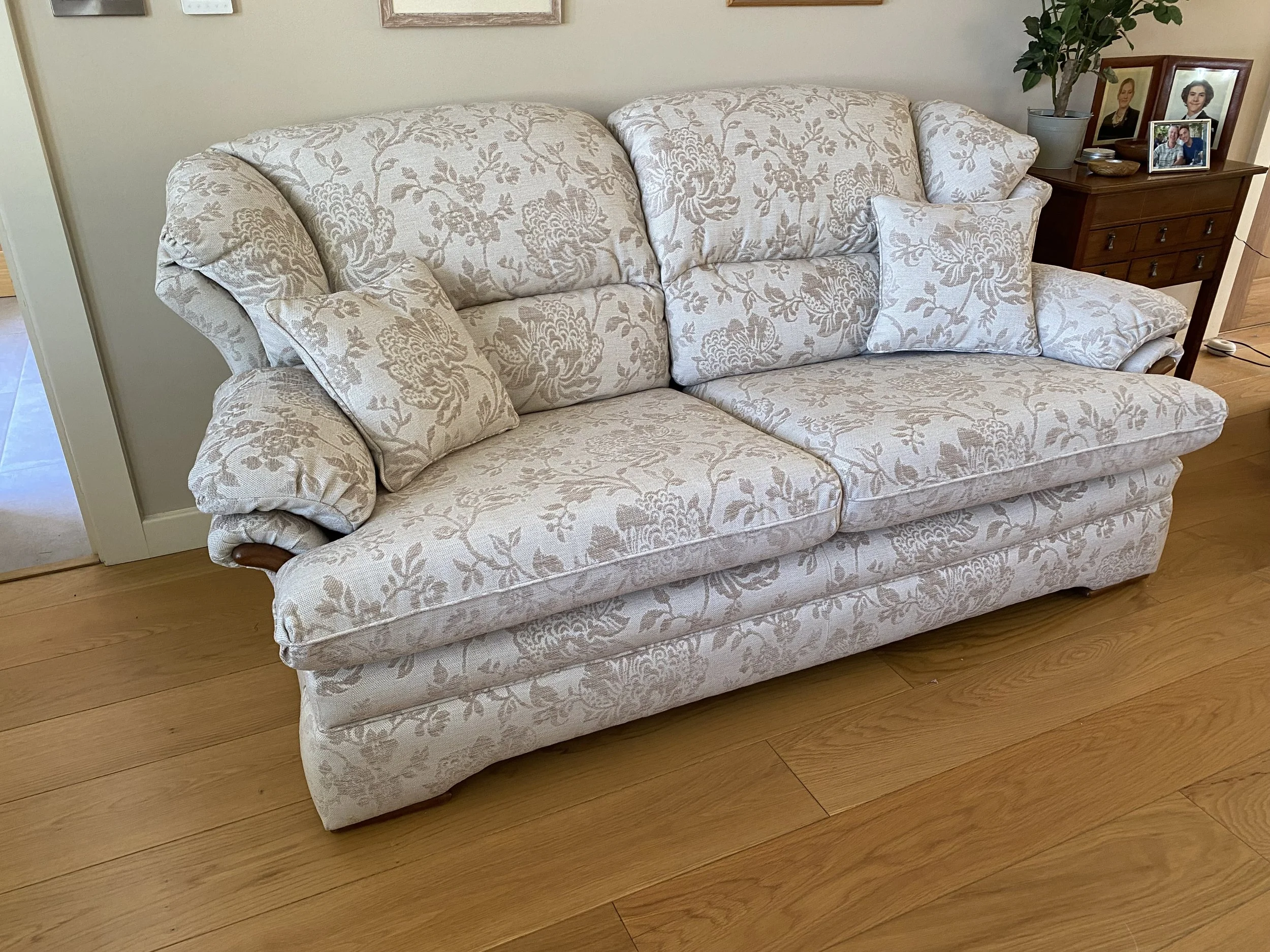 A floral patterned beige sofa with multiple cushions, positioned on a wooden floor, next to a wooden side table with family photos and a potted plant.