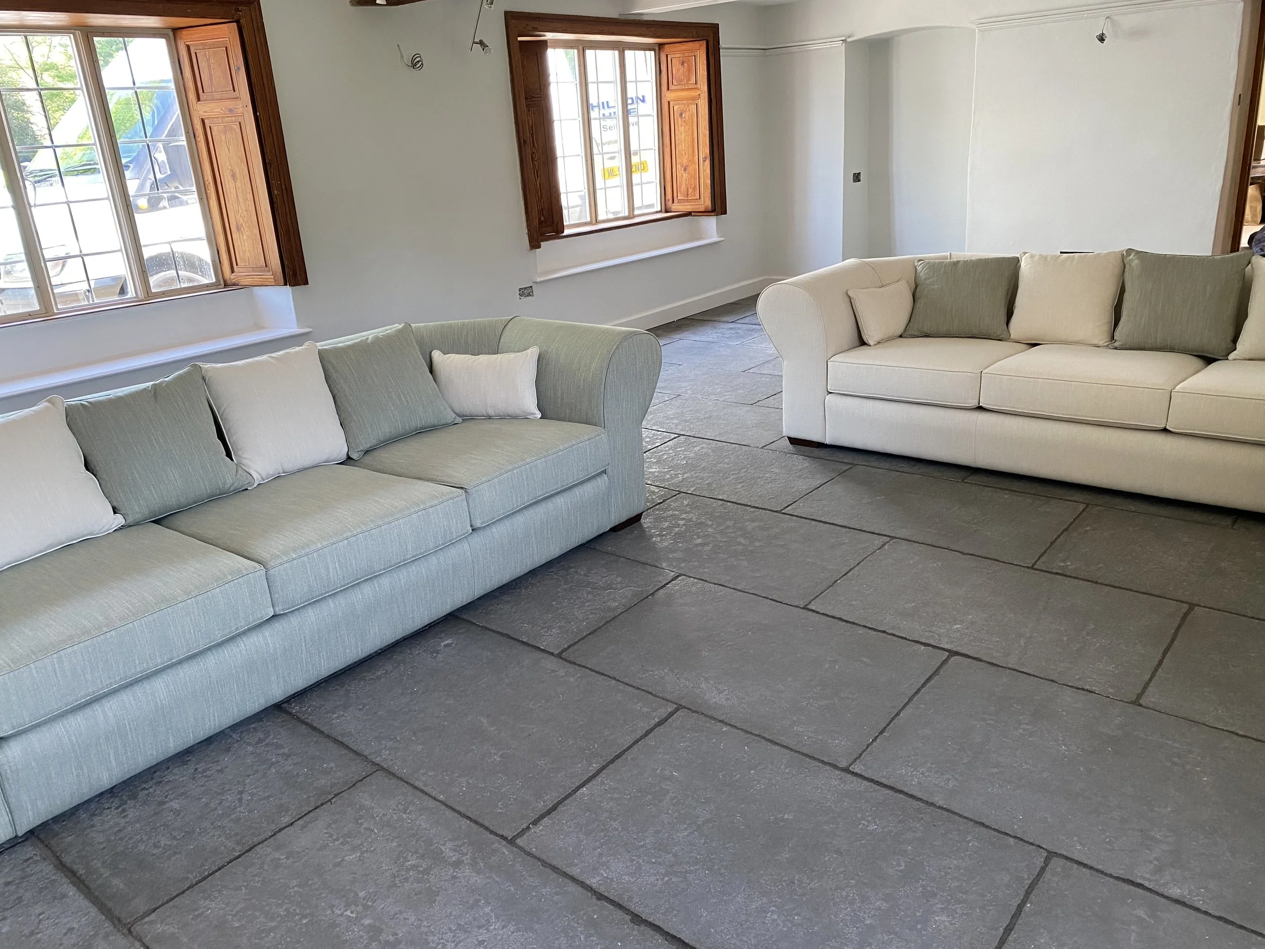 Living room with two large light-coloured sofas with cushions, large windows with wooden shutters, and a stone tile floor.