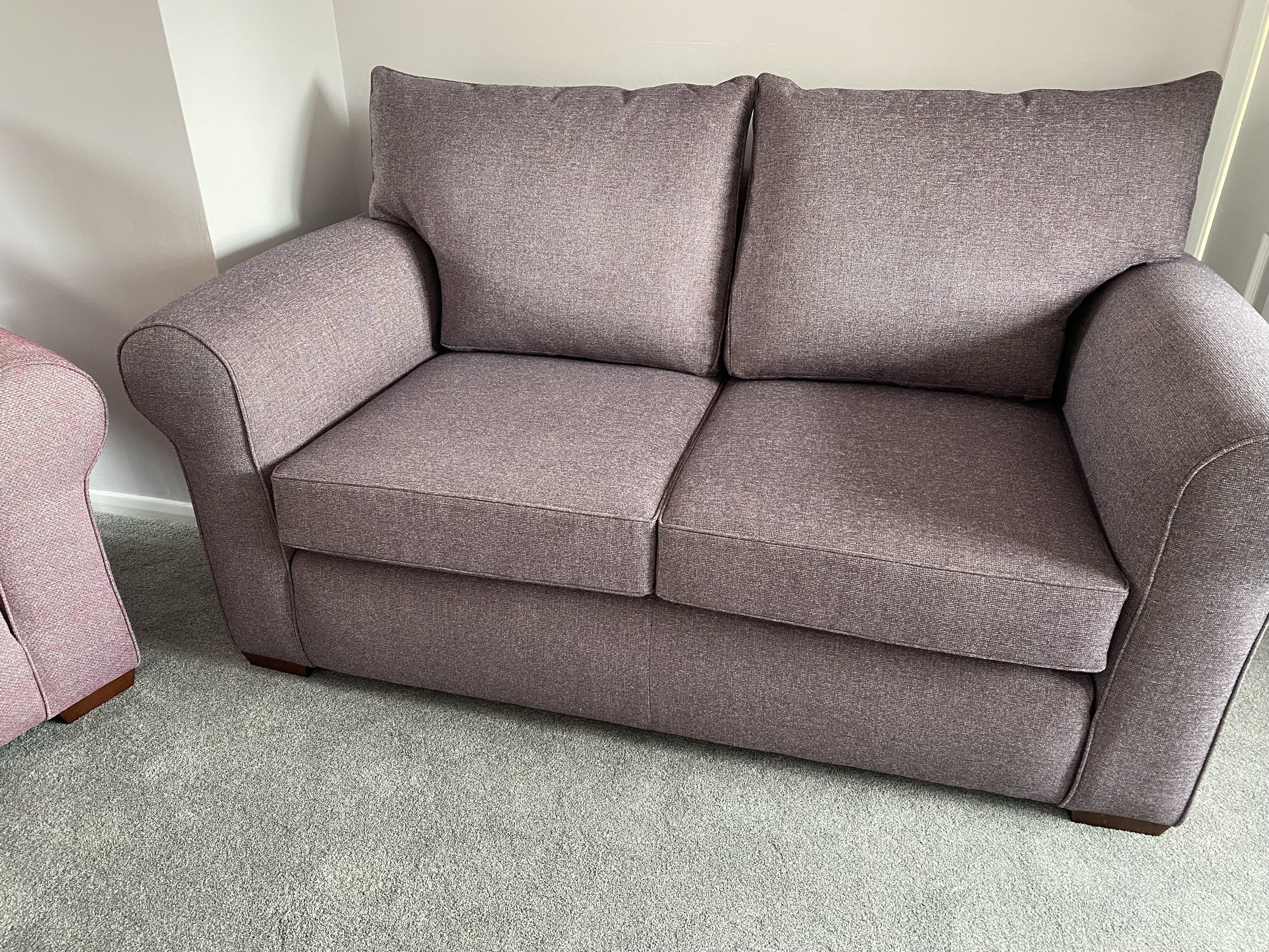 A purple upholstered loveseat with two cushions and rolled arms, placed on a beige carpeted floor next to a wall.