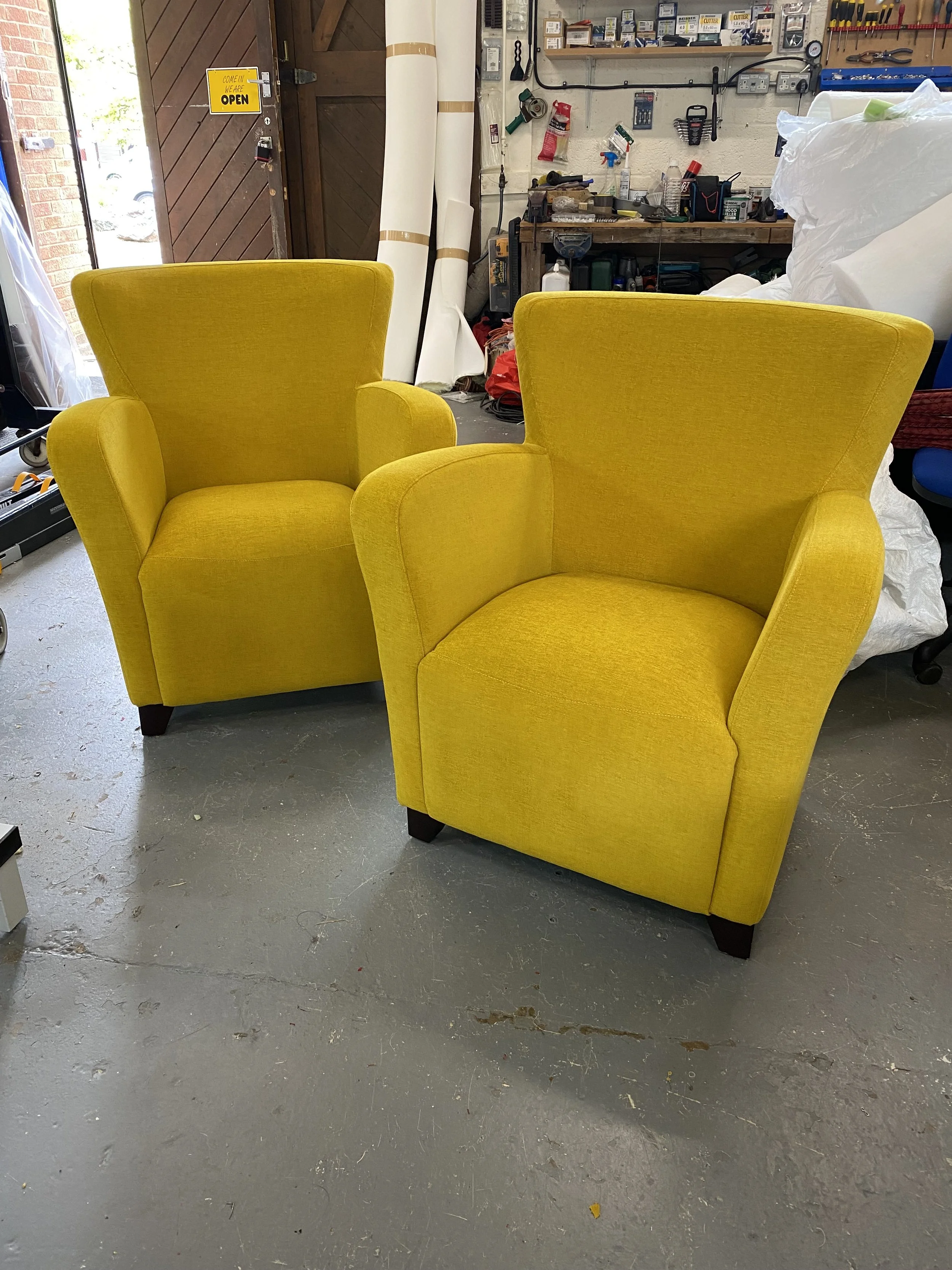 Two bright yellow upholstered armchairs in a workshop or storage area with tools and supplies on the shelves behind.