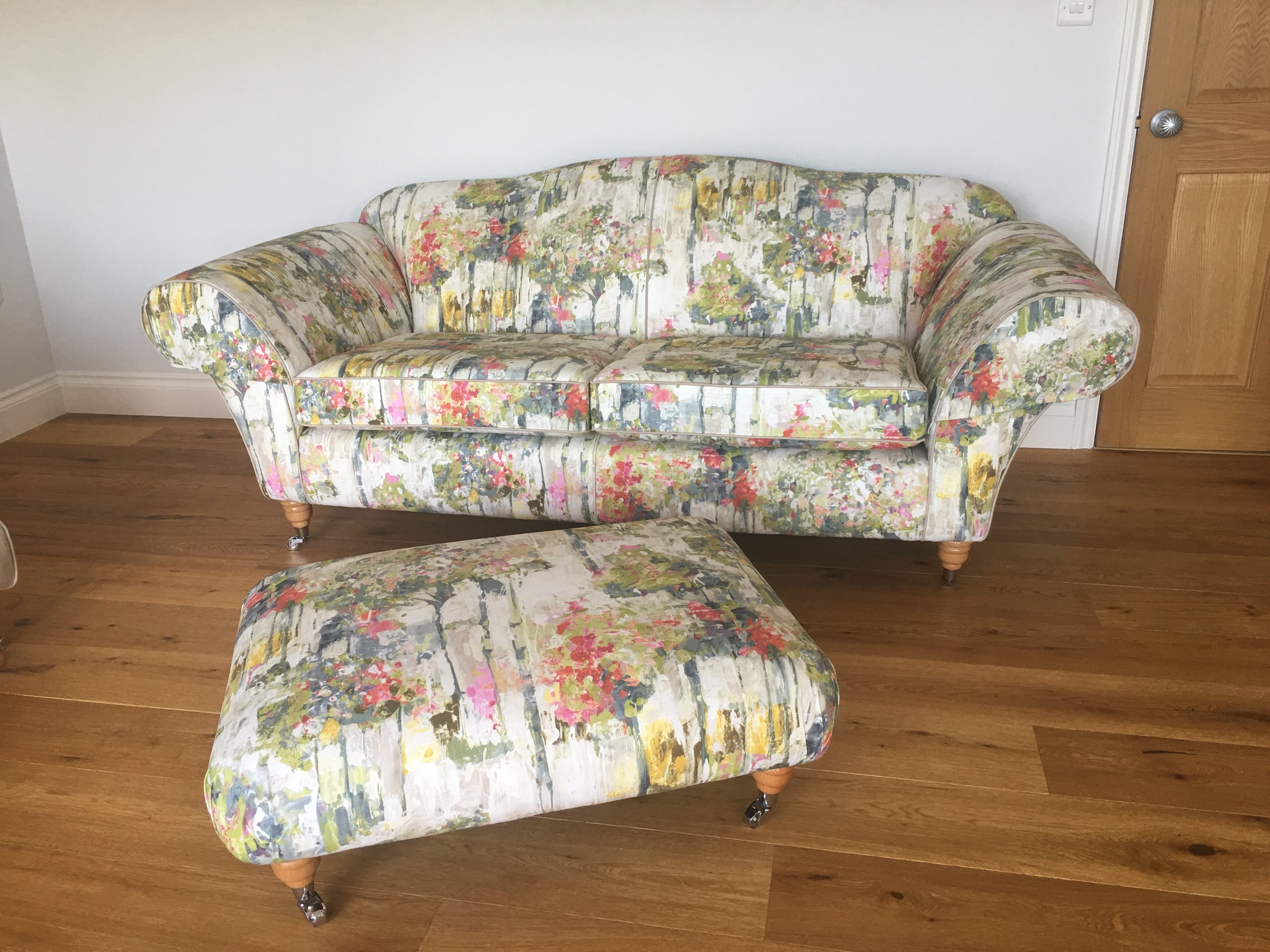 A floral upholstered sofa with matching floral ottoman on wooden floor in a room with white walls and a wooden door.
