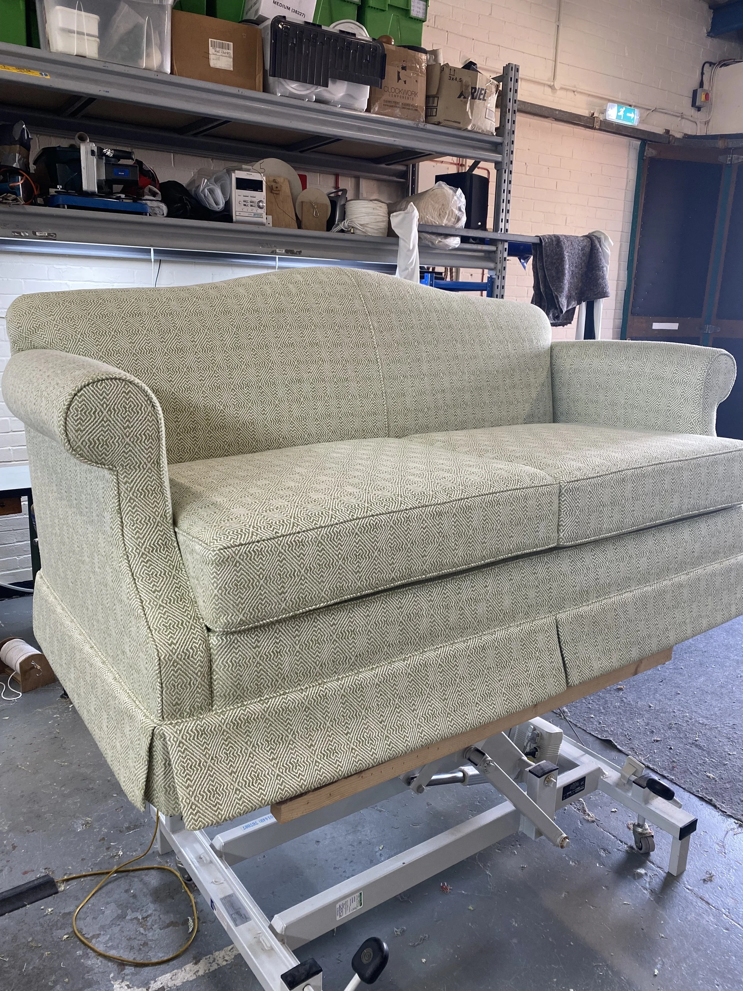 A beige upholstered sofa resting on a medical or dental examination table in a warehouse or workshop setting.