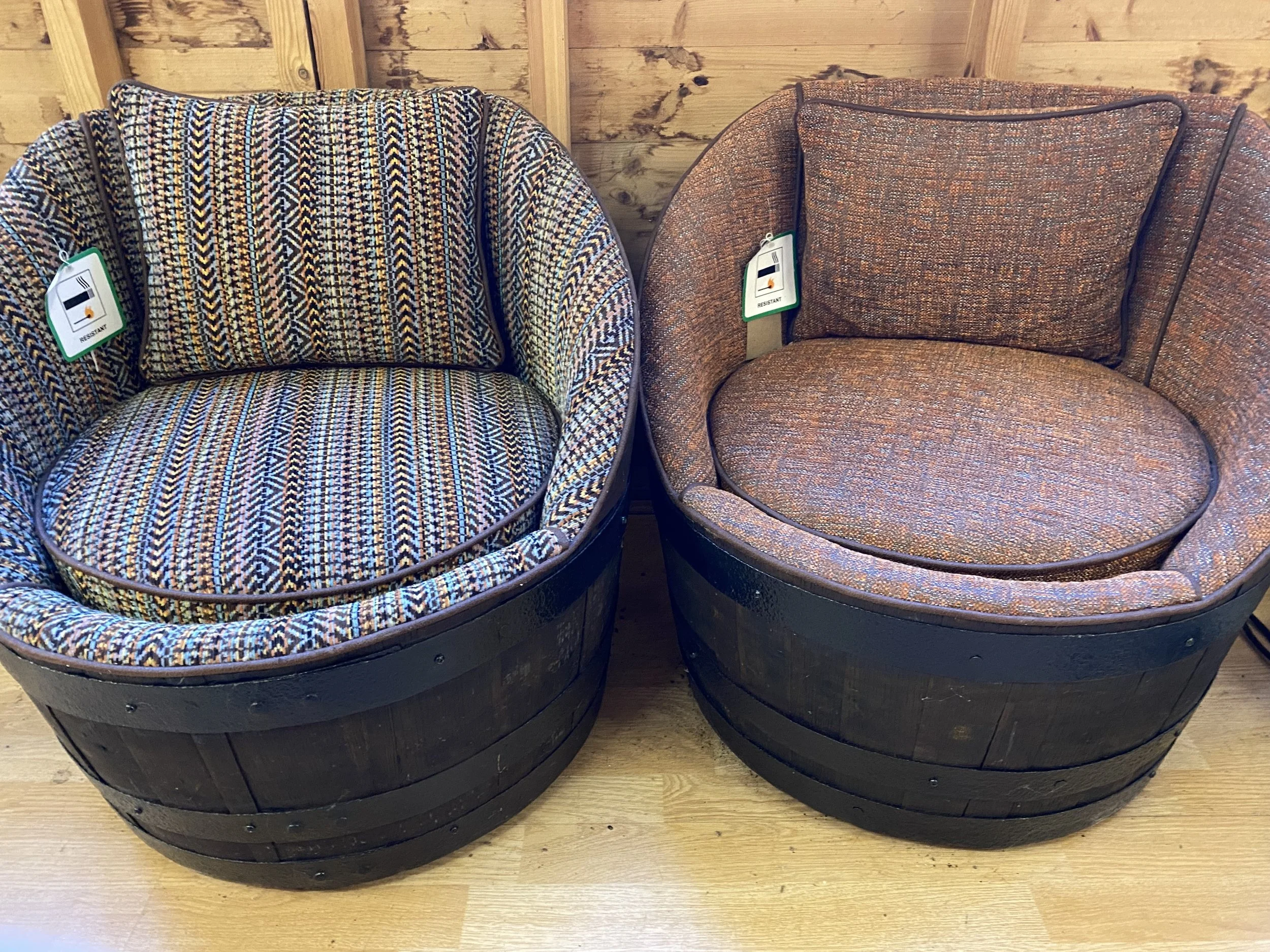 Two barrel-shaped chairs with patterned fabric upholstery, placed side by side on a wooden floor against a wooden wall, each with a tag indicating they are resistant.