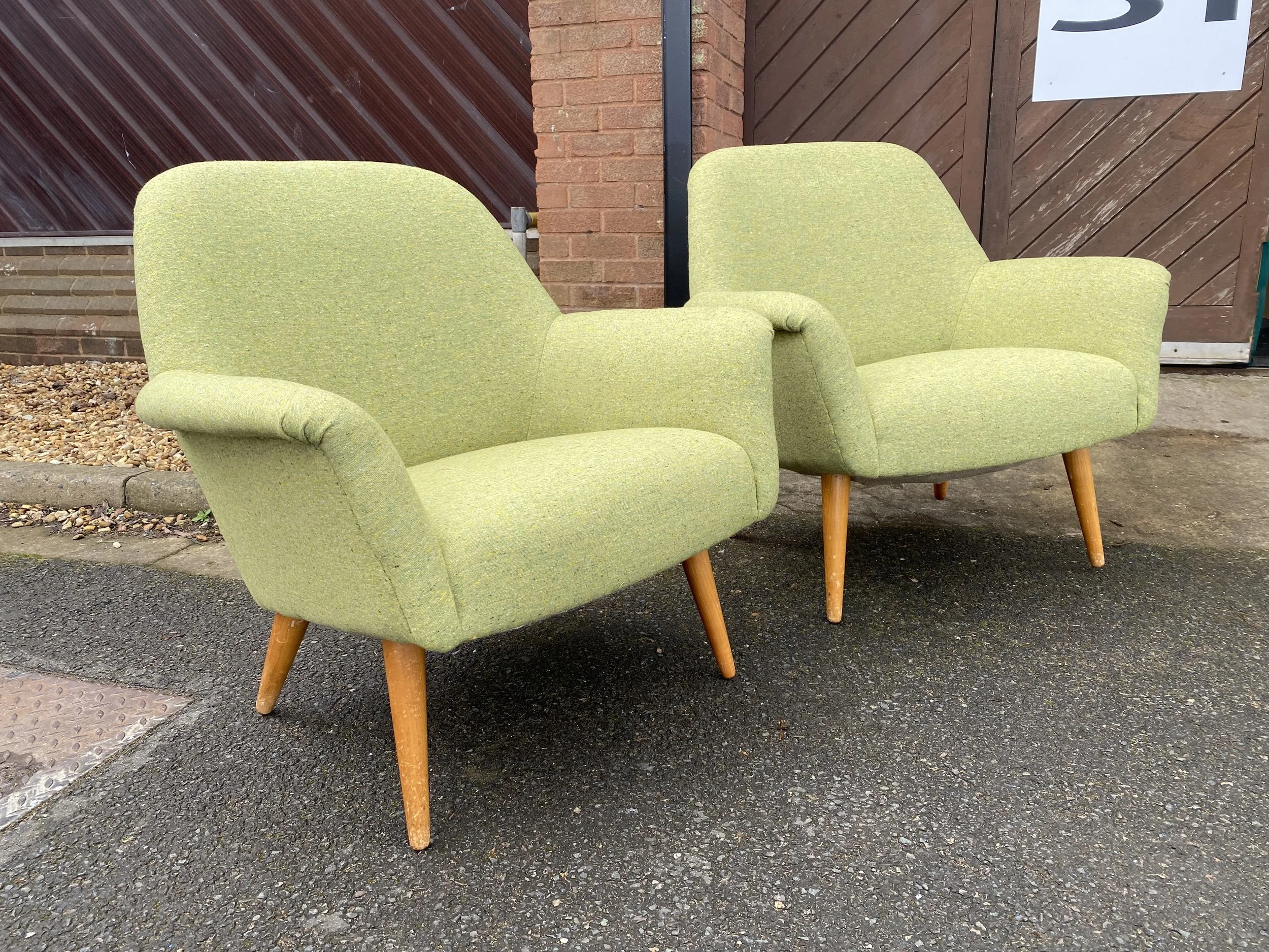Two vintage-style green upholstered armchairs with wooden legs outside on a paved area near brick and wood walls.