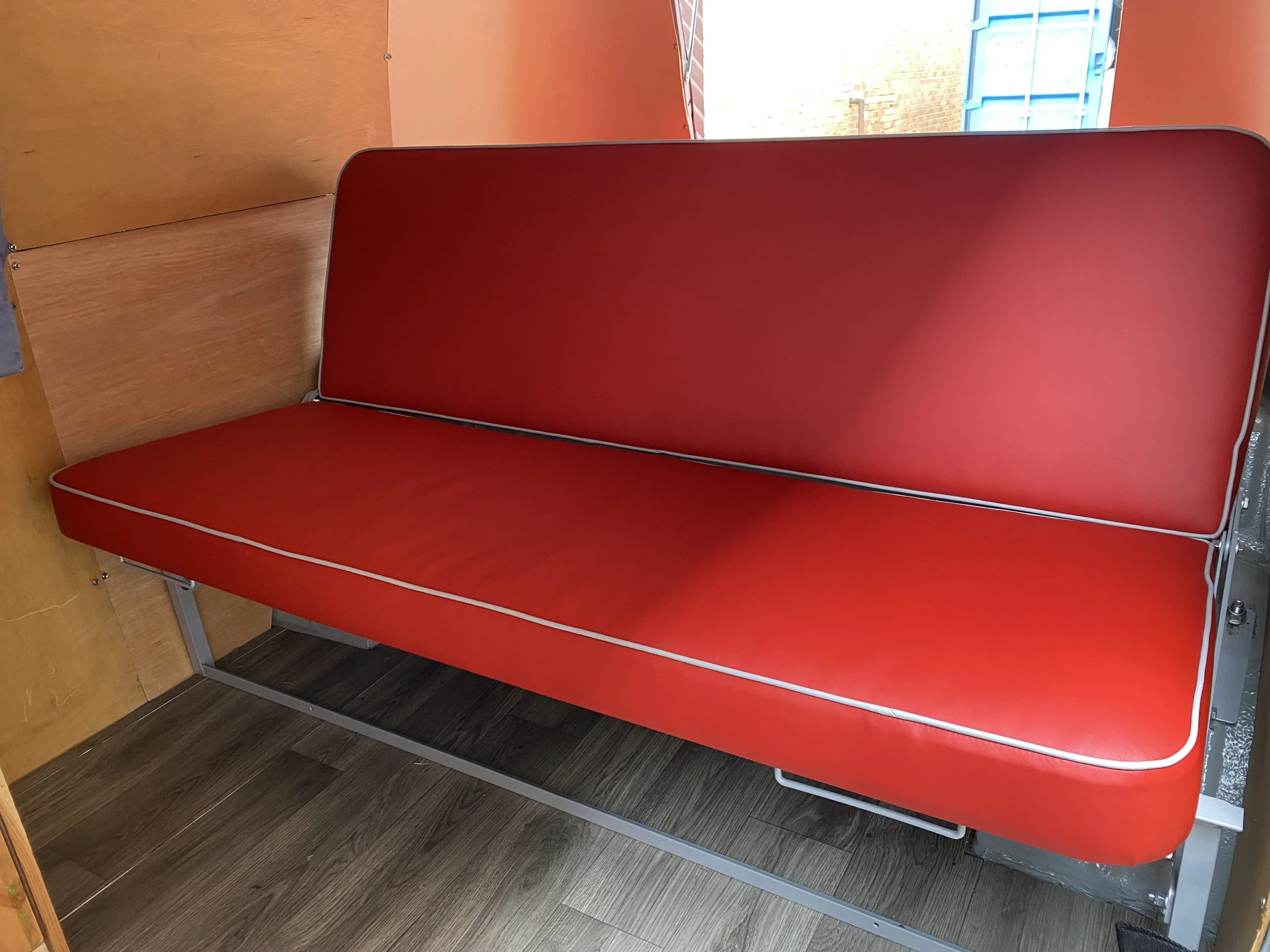 Red rock’n’roll bench seating in campervan with metal frame in a room with wooden wall panels and window showing brick building outside.