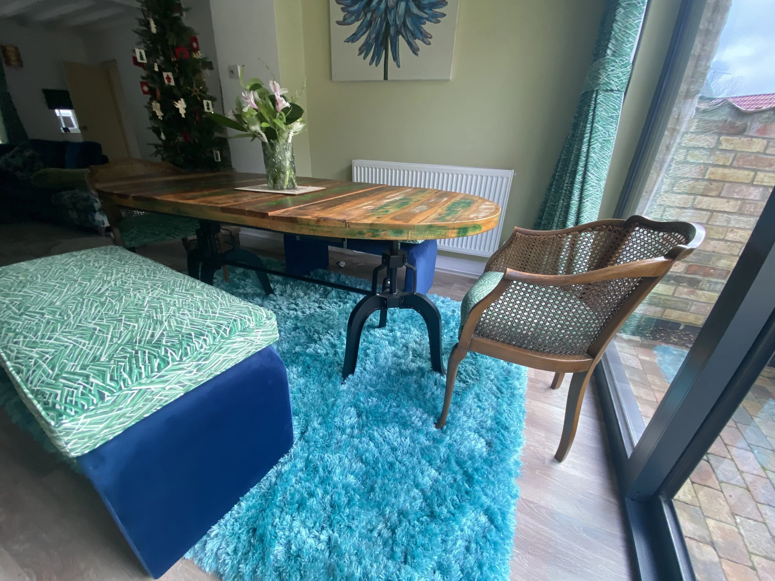A cozy dining area with a wooden table, two woven cane chairs, a turquoise shaggy rug, a vase of pink lilies on the table, and a view of a brick patio outside through a glass sliding door.