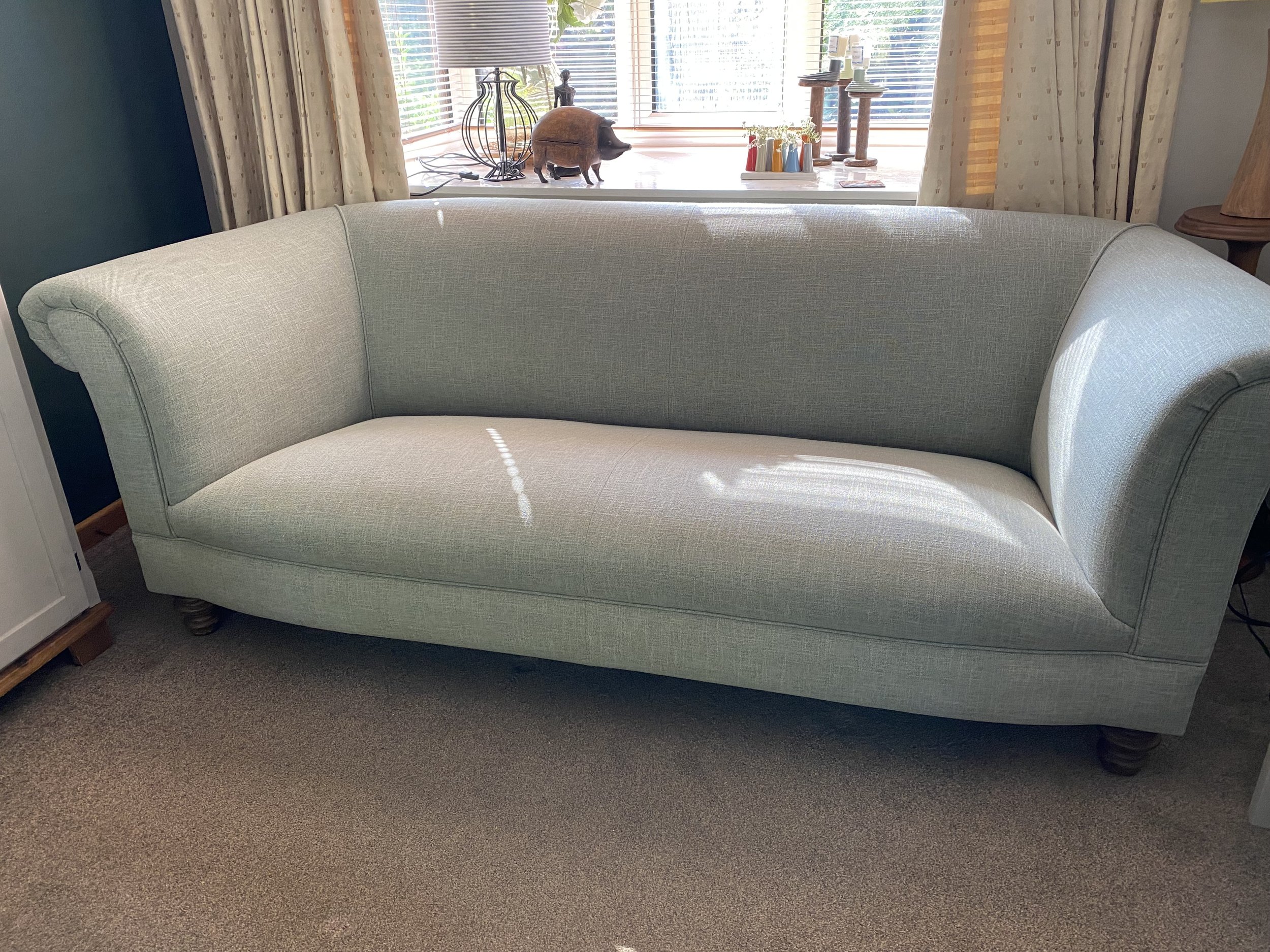 Light green upholstered sofa in a living room with a window behind, decorated with curtains, small wooden feet, and sunlight reflections.
