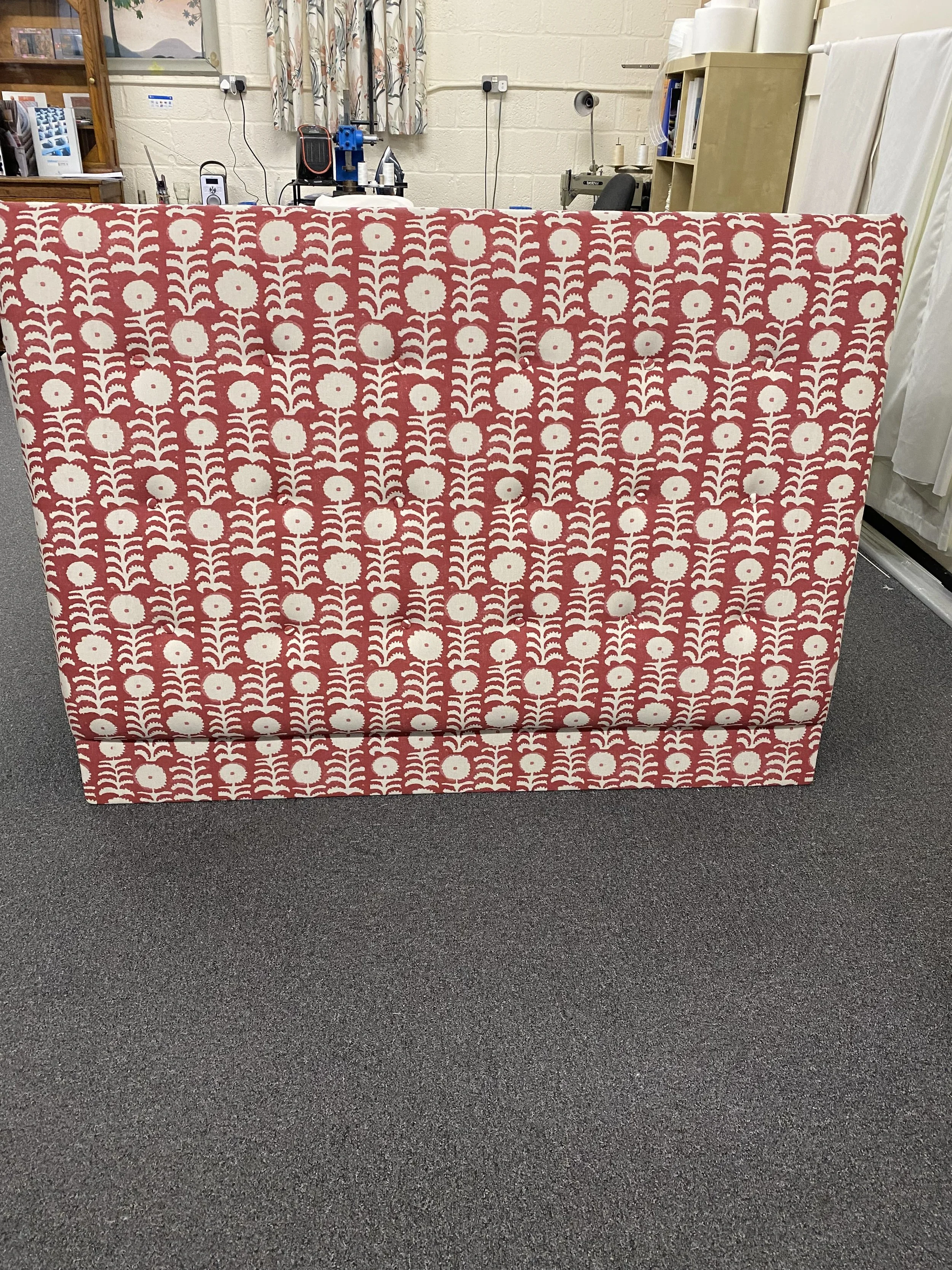 Red and white upholstered headboard with a floral pattern on a gray carpeted floor in a workshop or showroom.