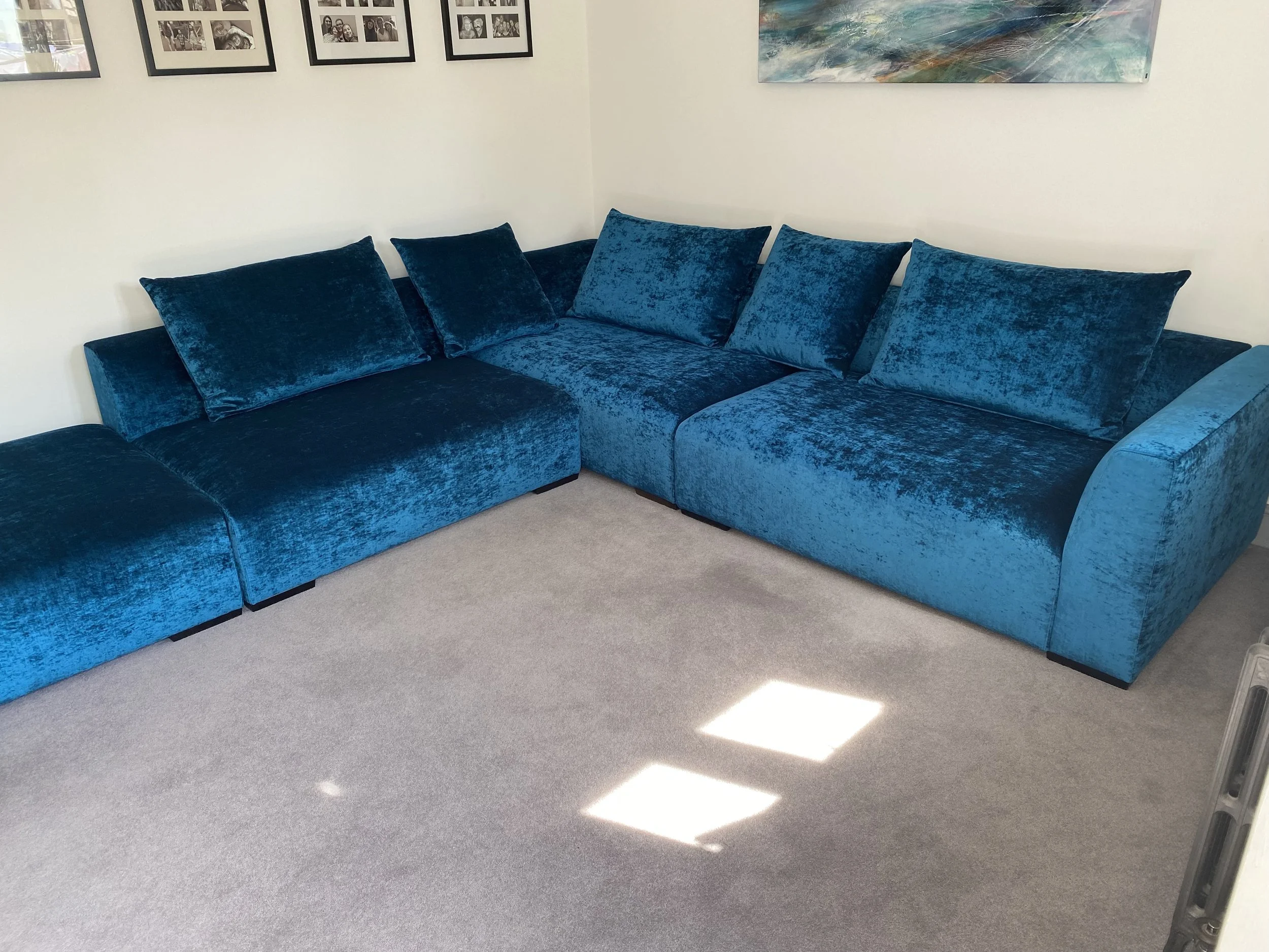 A blue velvet sectional sofa with five matching cushions in a living room with beige carpet and framed photos on a white wall.
