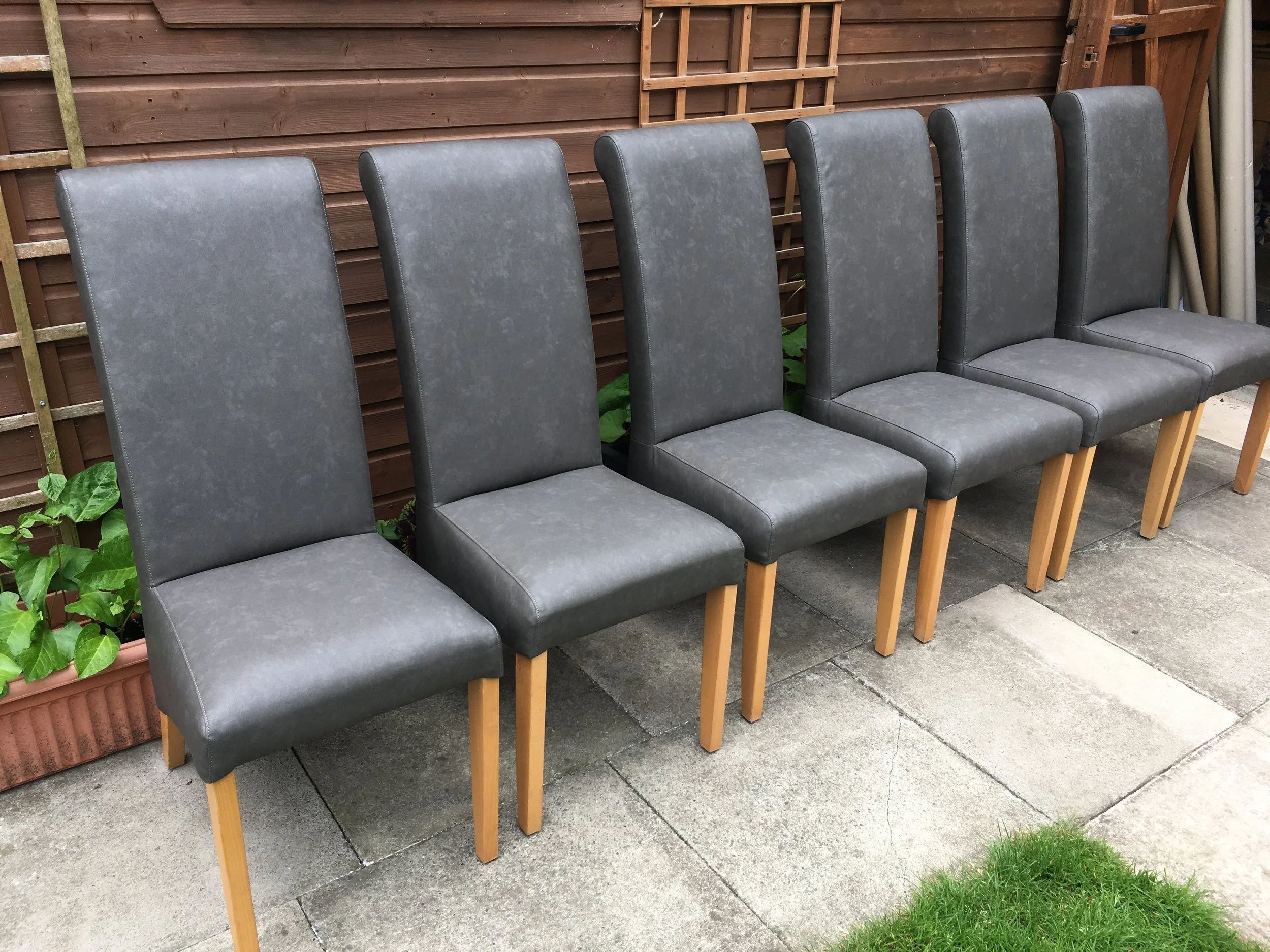 Six gray upholstered chairs with wooden legs arranged in a row outdoors on a concrete patio next to a wooden fence and some green plants.