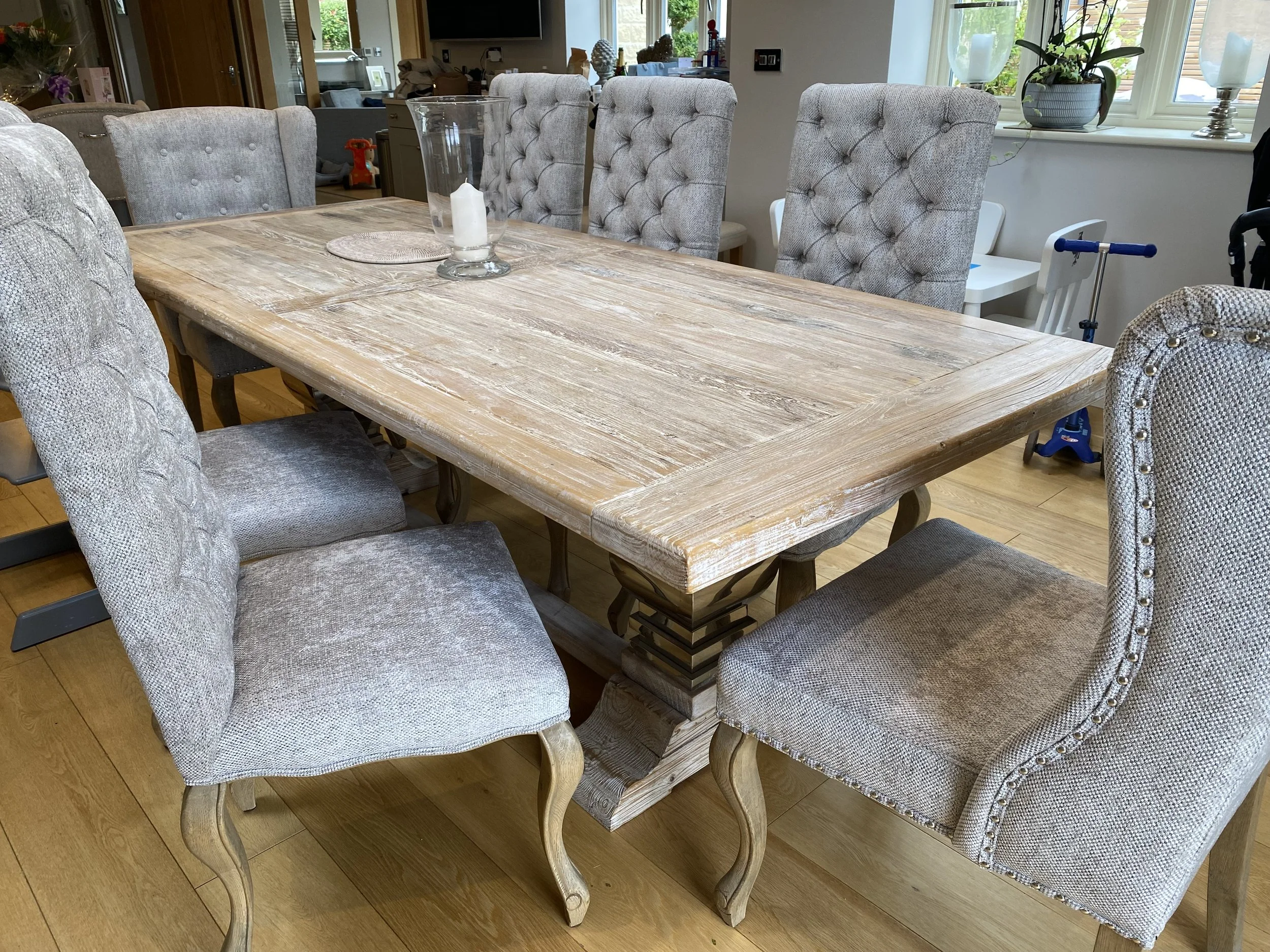 A dining room table with eight upholstered chairs, a glass hurricane candle holder with a white candle, and a round beige placemat, with a window in the background showing plants and a view outside.