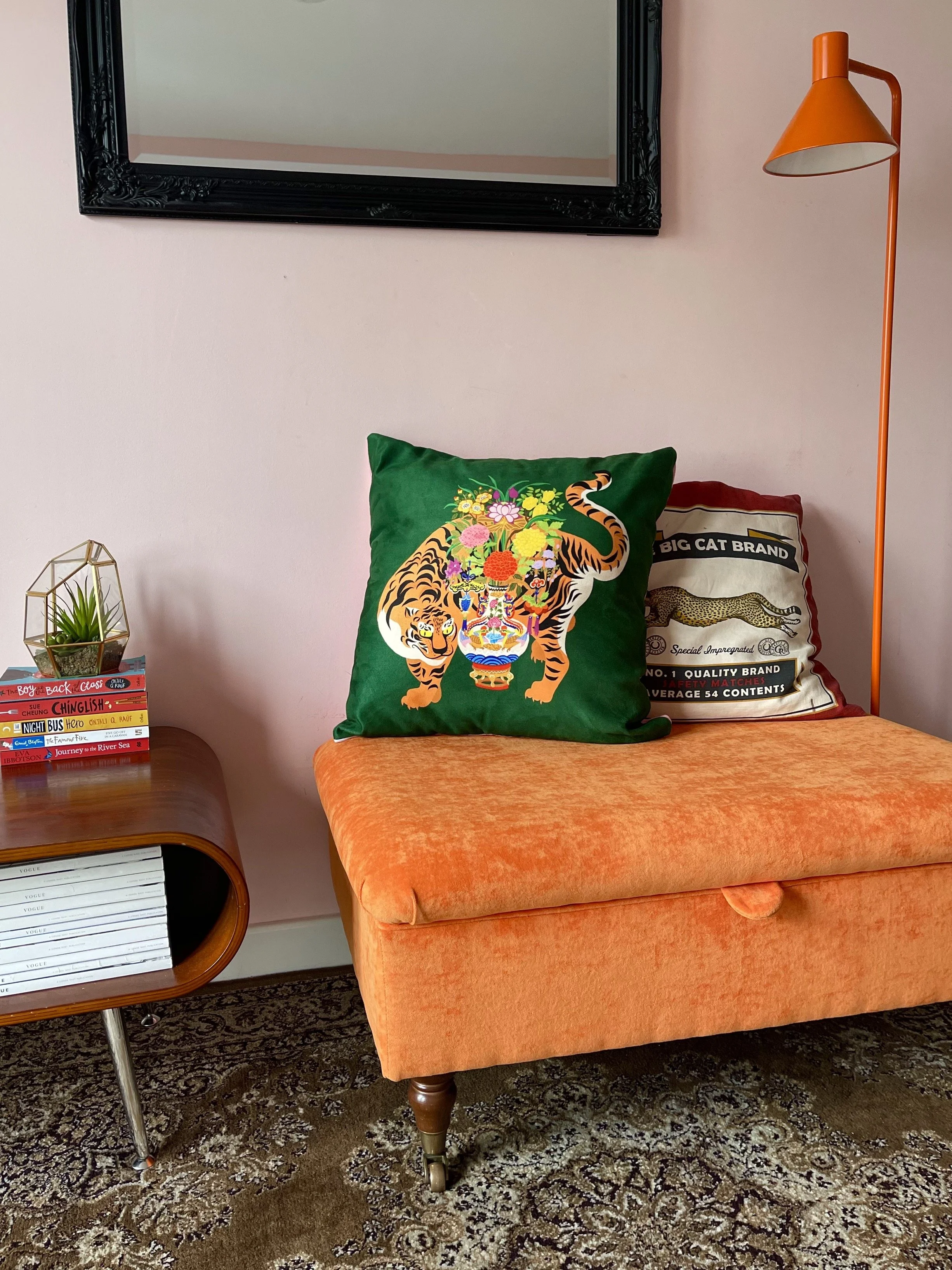 A living room with a pink wall, a black-framed mirror, an orange floor lamp, a vibrant orange upholstered storage footstool with decorative pillows on top, a small side table with a plant and a stack of books, and a patterned rug.