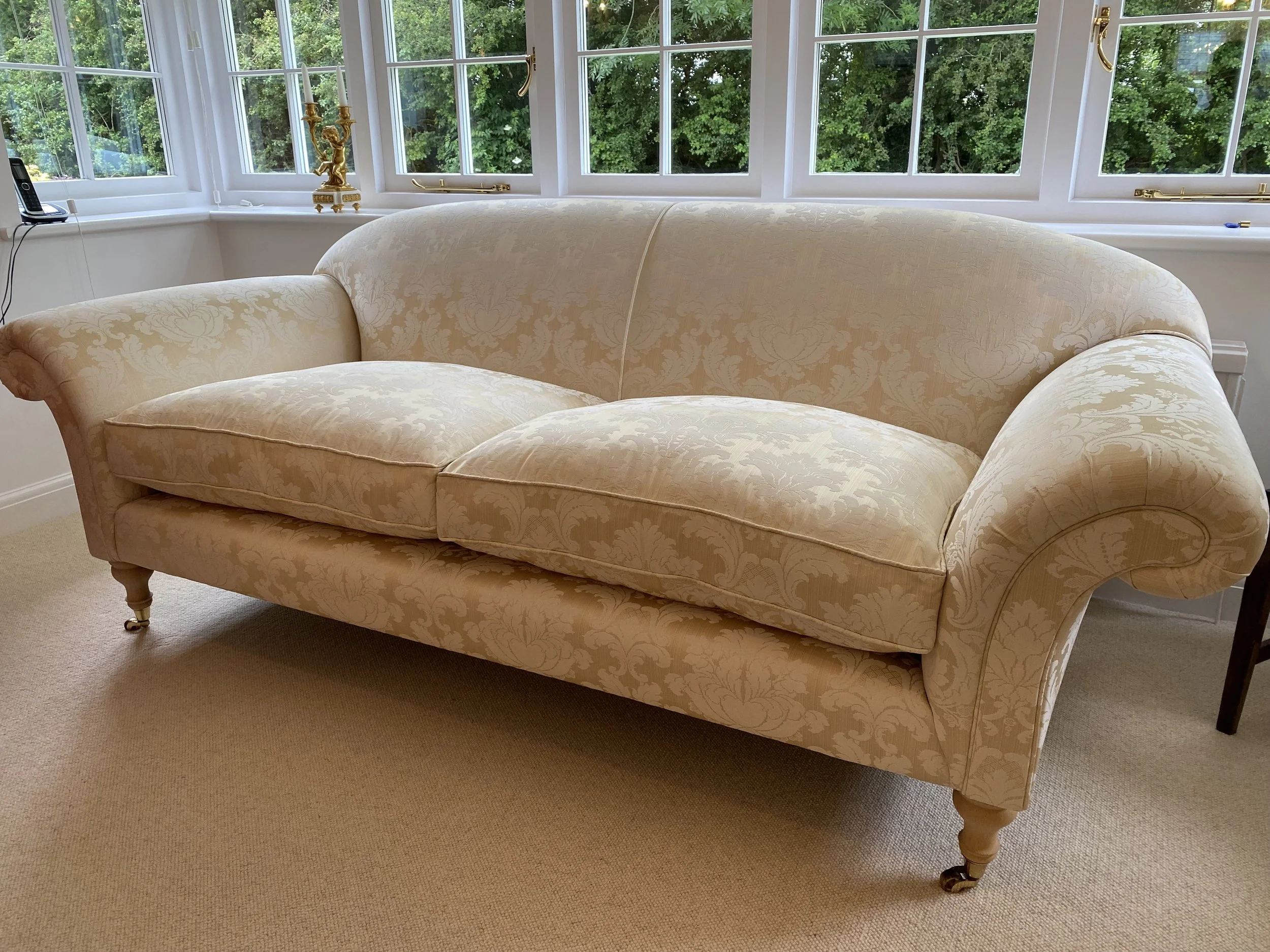 Elegant cream-colored sofa with floral pattern in front of large windows showing green foliage outside.