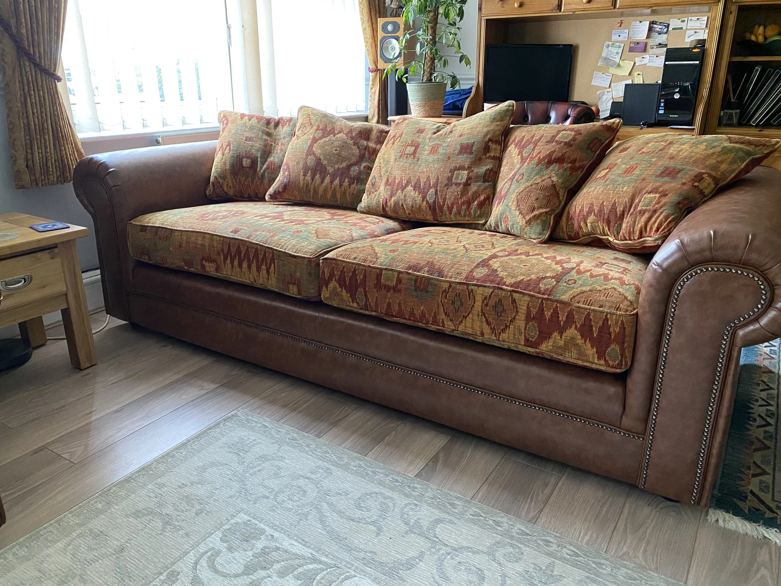 A brown leather sofa with colorful, patterned cushions in a living room near a window with curtains and a small wooden side table.
